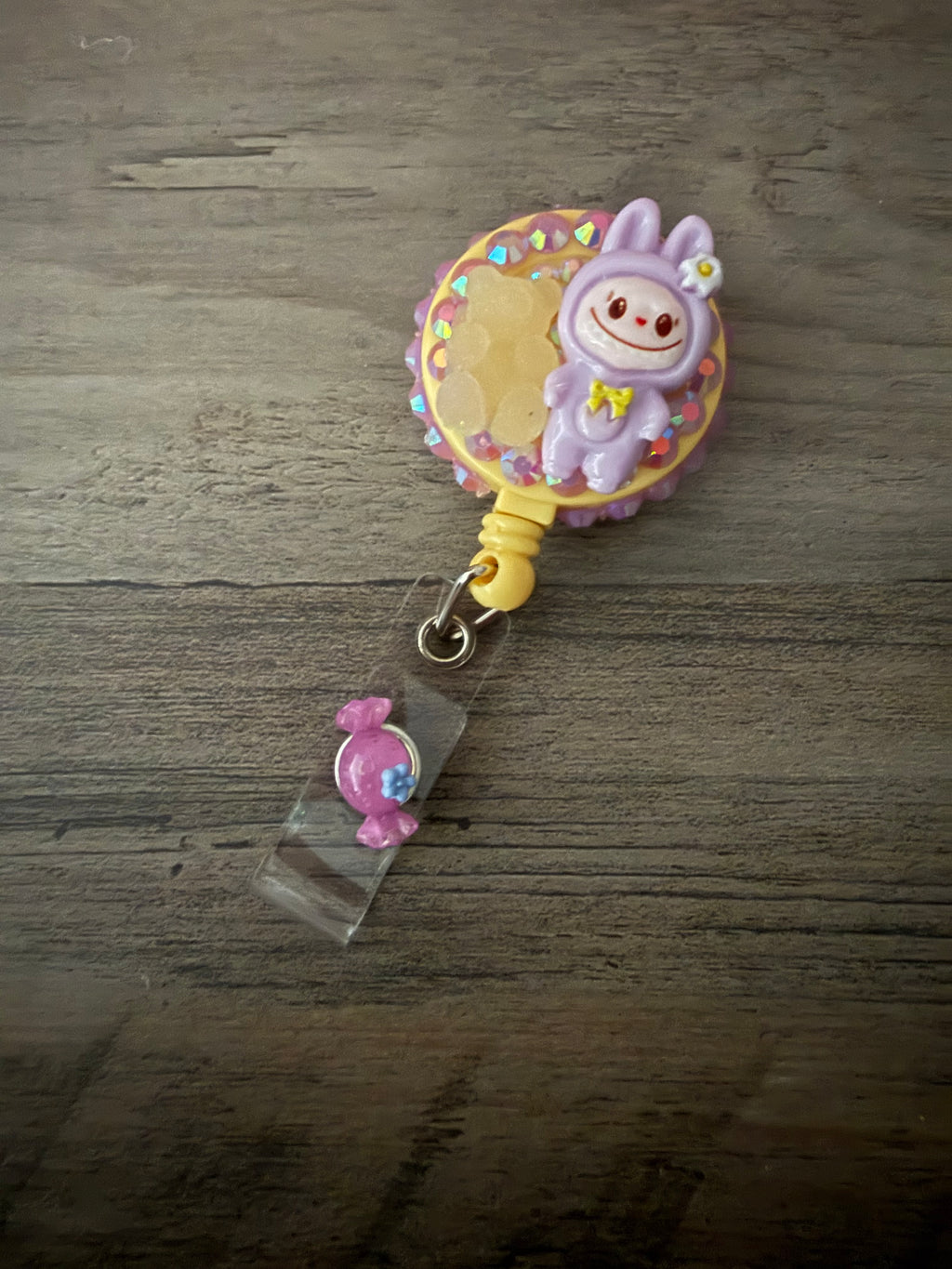 A handcrafted, pastel purple monster in a hooded outfit on a yellow retractable badge reel, decorated with pink rhinestones, a gummy bear charm, and a small pink candy charm on the strap.