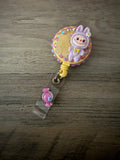 A handcrafted, pastel purple monster in a hooded outfit on a yellow retractable badge reel, decorated with pink rhinestones, a gummy bear charm, and a small pink candy charm on the strap.