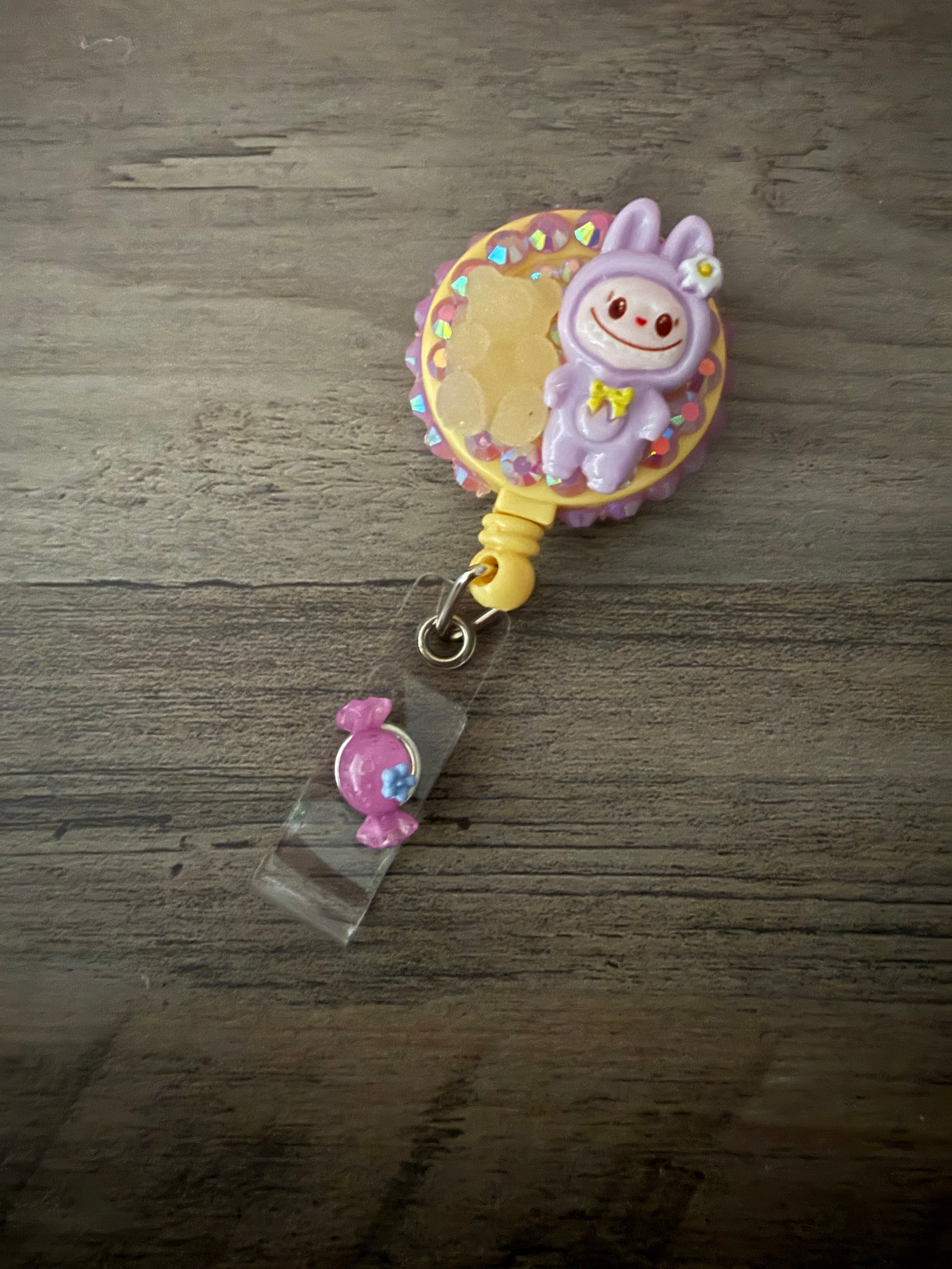 A handcrafted, pastel purple monster in a hooded outfit on a yellow retractable badge reel, decorated with pink rhinestones, a gummy bear charm, and a small pink candy charm on the strap.