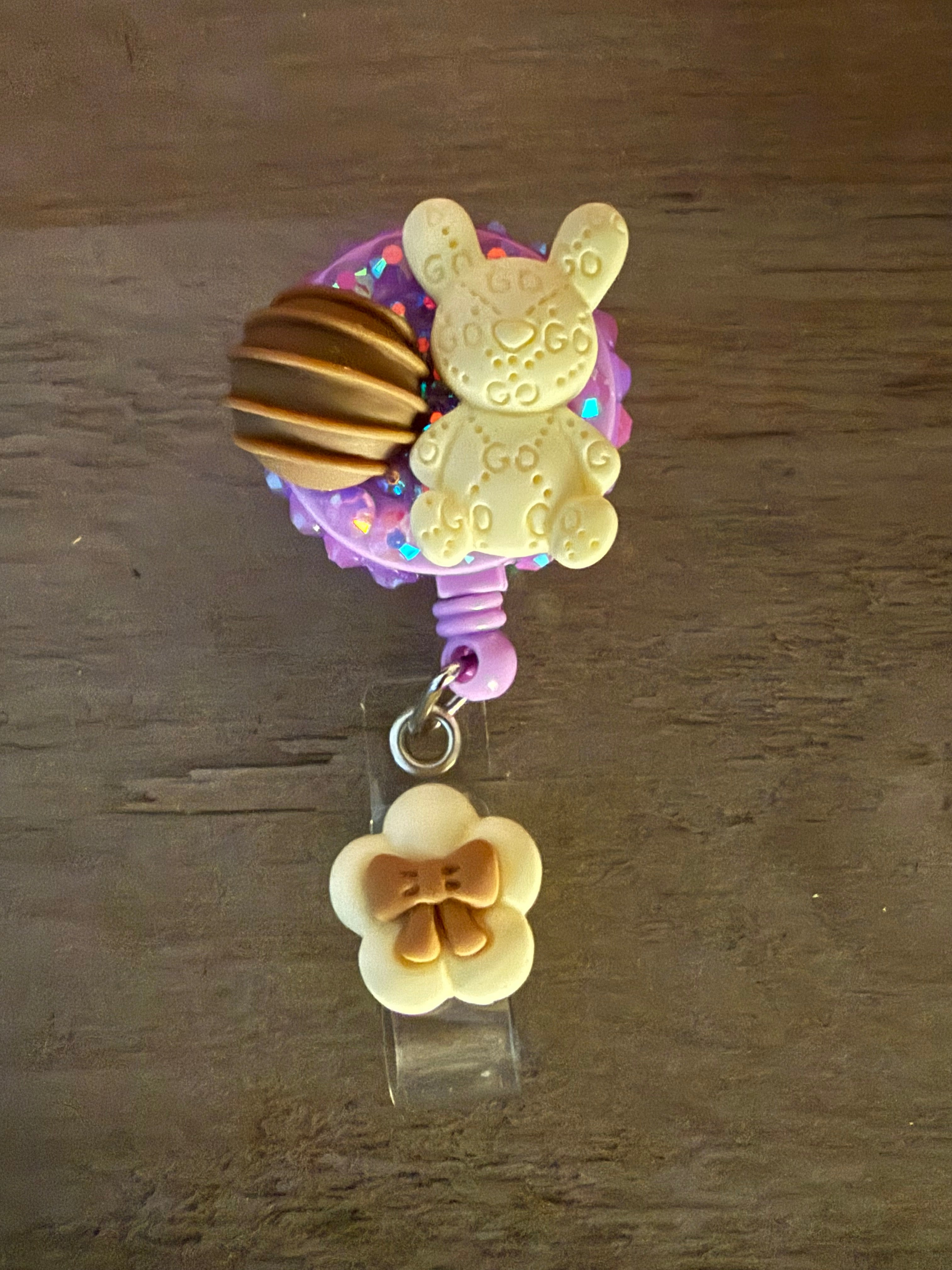A retractable badge reel featuring a decoden-style purple glitter base decorated with a designer-patterned bunny charm and a chocolate truffle, with a white flower charm featuring a brown bow hanging from the clip.