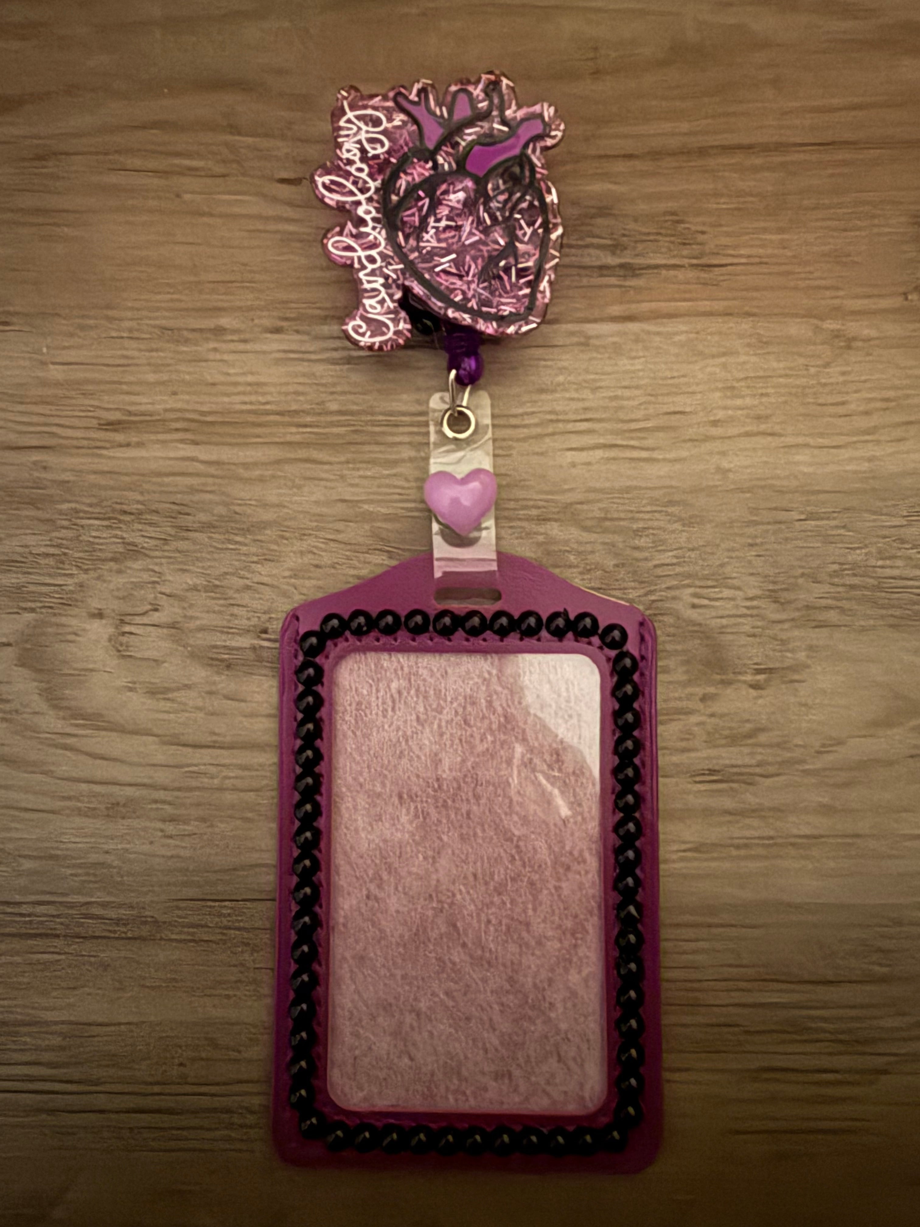 A purple ID badge holder with black beaded trim, featuring a glittery cardiology-themed heart badge reel and a light purple heart accent.