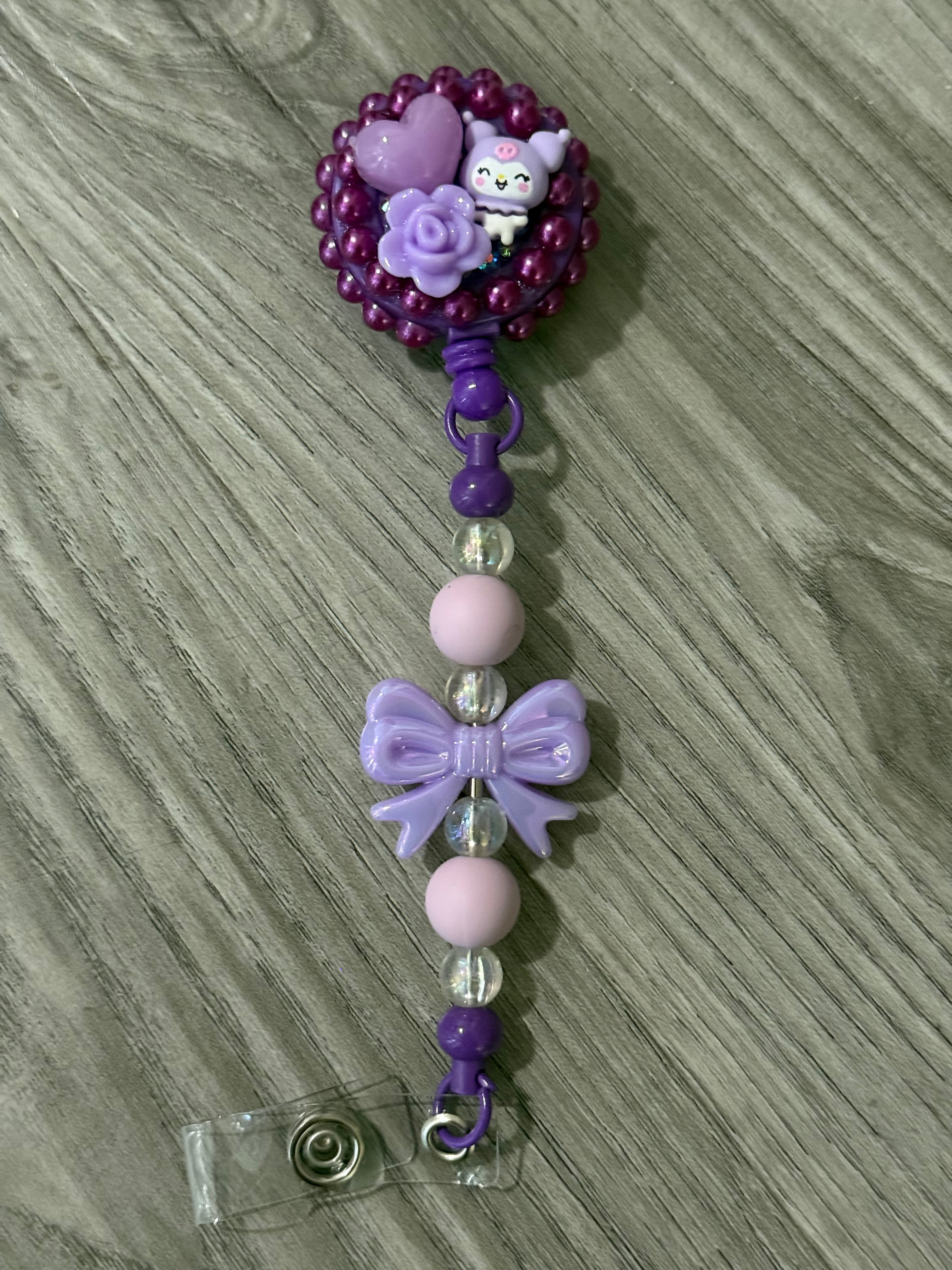 Cute badge reel featuring a whimsical design with a purple top adorned with a kuromi inspired character, a heart, and a rose, complemented by a decorative purple bow and colorful beads on the strap.