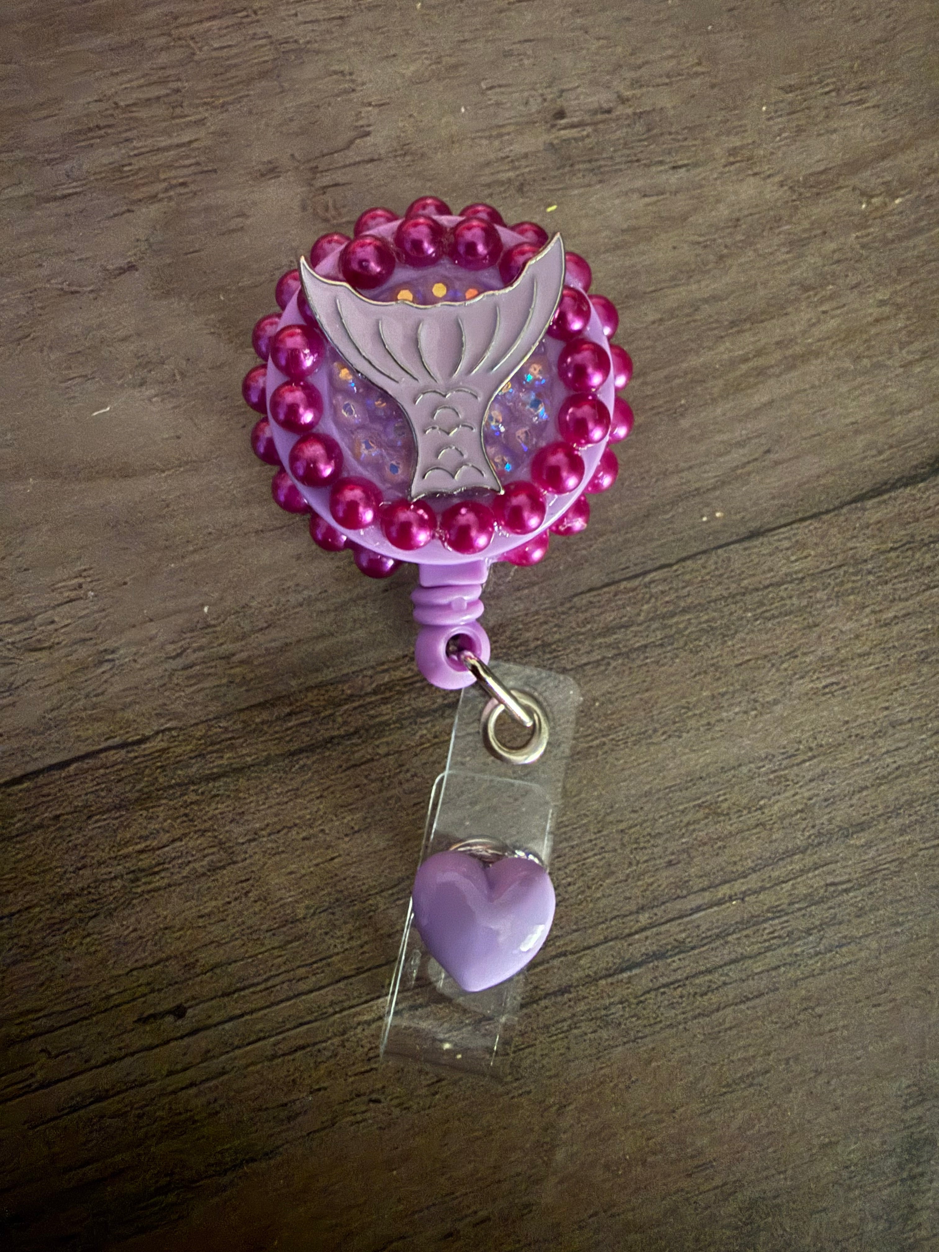 Handmade purple badge reel decorated with dark pink pearls, a purple mermaid tail charm, and iridescent glitter, featuring a purple heart dangle.