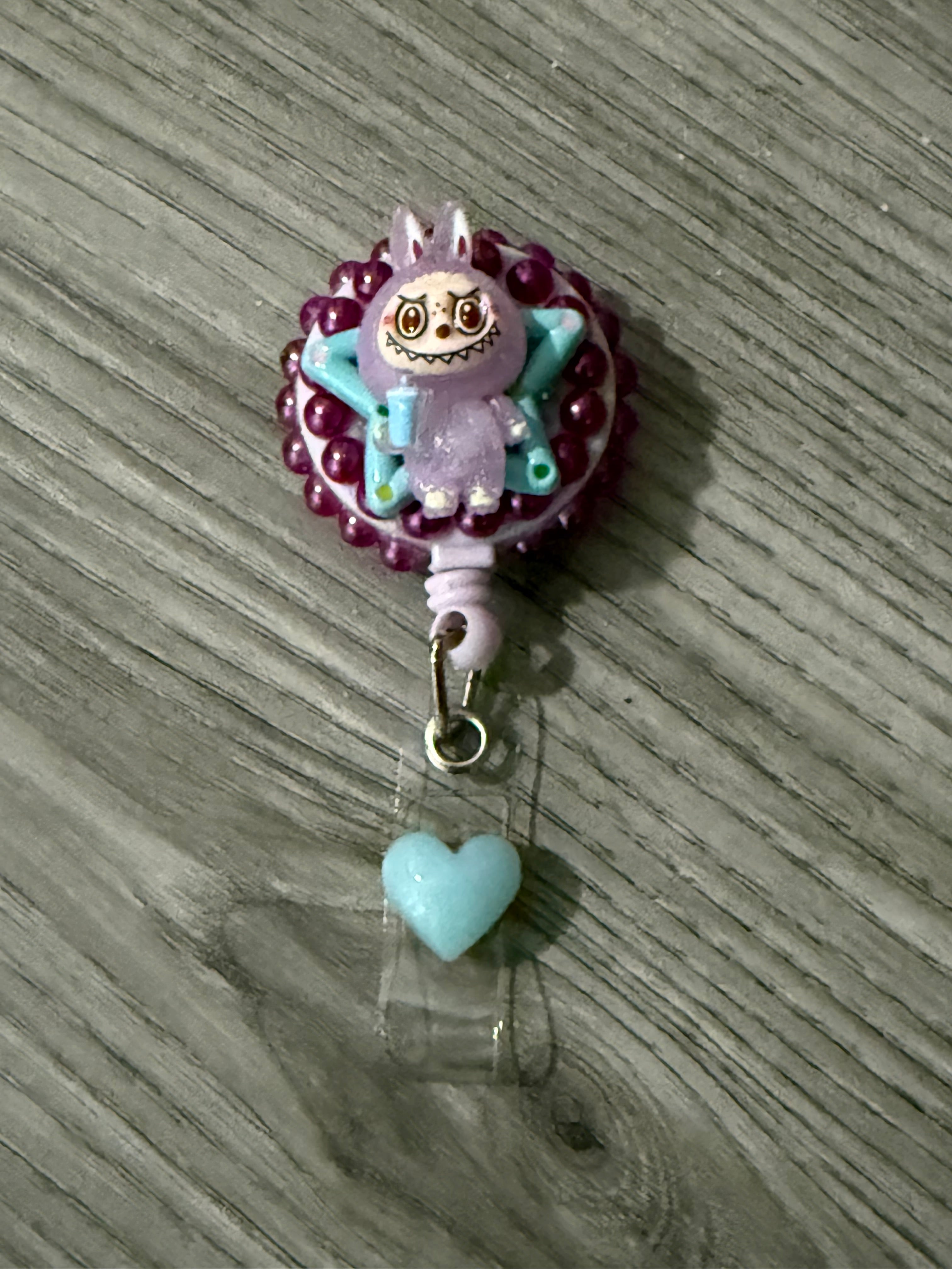 Cute badge reel featuring a whimsical purple monster with bunny ears and a mischievous grin, surrounded by purple beads and a blue heart charm.