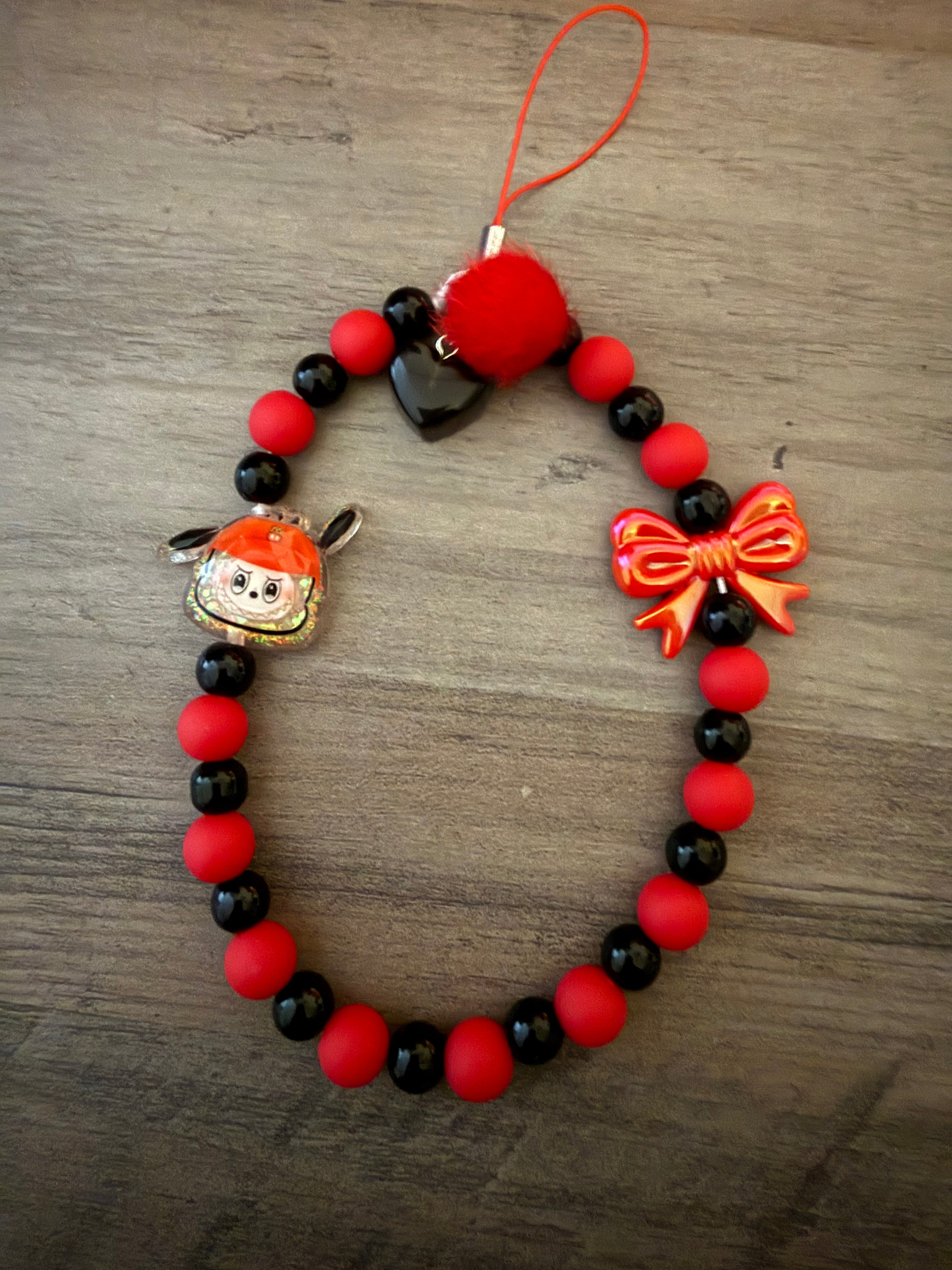 A beaded phone charm featuring matte red and shiny black beads, a red pom-pom, a metallic red bow, a black heart charm, and a small monster charm in a red hat on a wooden surface.