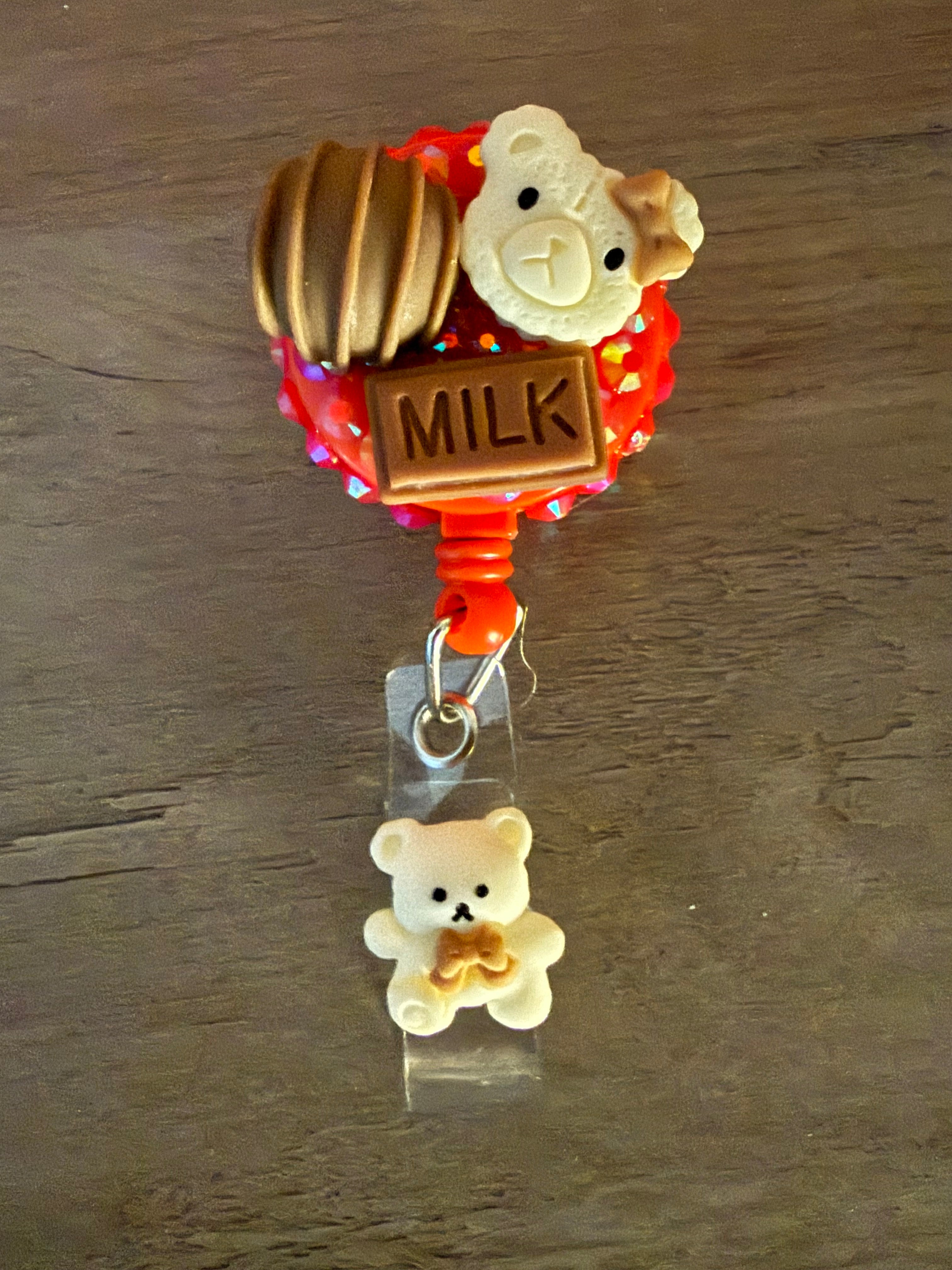 Handmade red glitter badge reel decorated with a bear-shaped cookie, a chocolate truffle, and a milk chocolate bar charm, finished with a small white bear dangle.