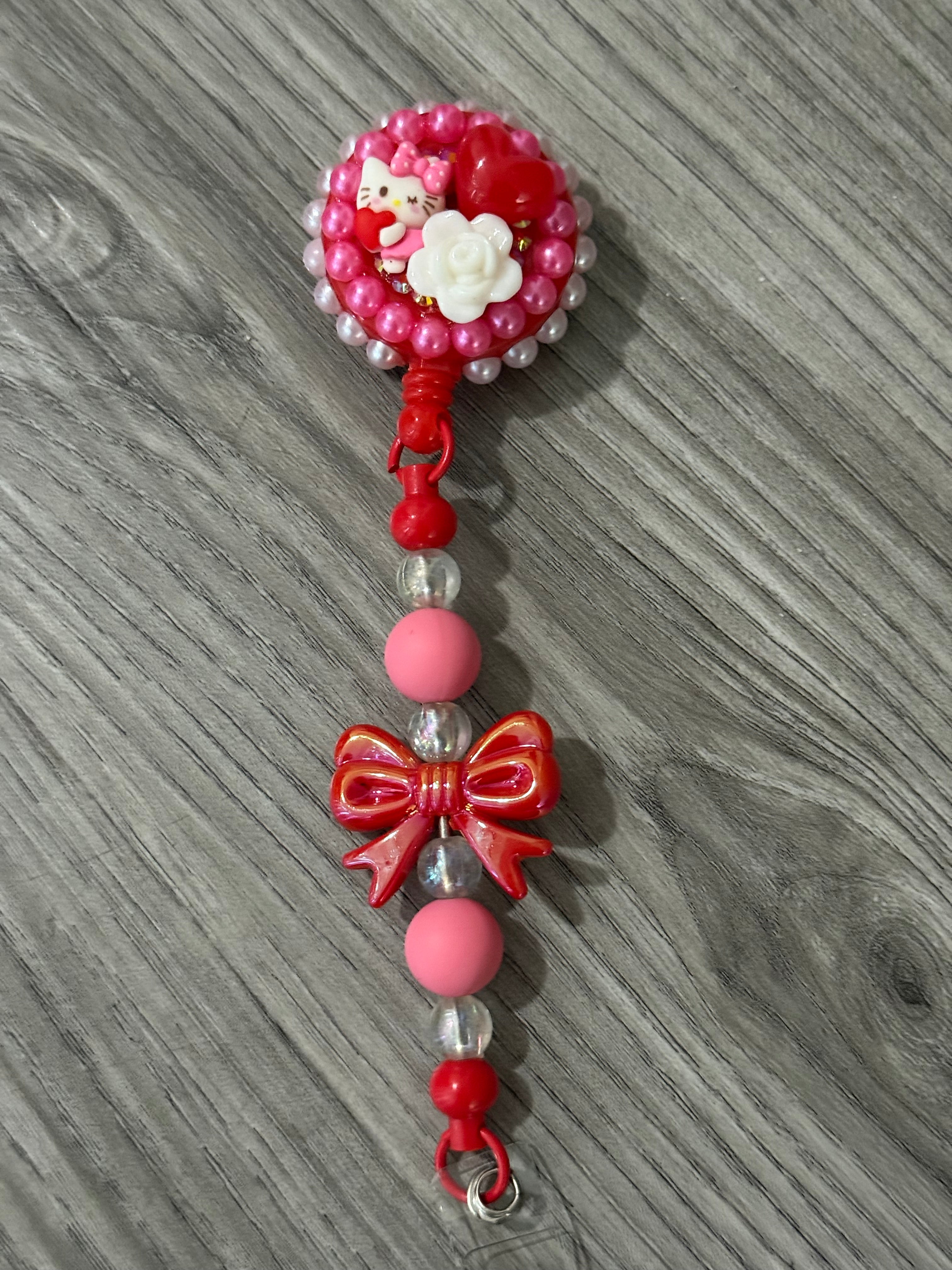 Cute badge reel featuring a playful design with a pink and white Hello Kitty inspired character, a red heart, and a white rose, surrounded by pink pearl-like beads, complemented by a decorative red bow and colorful beads on the strap.
