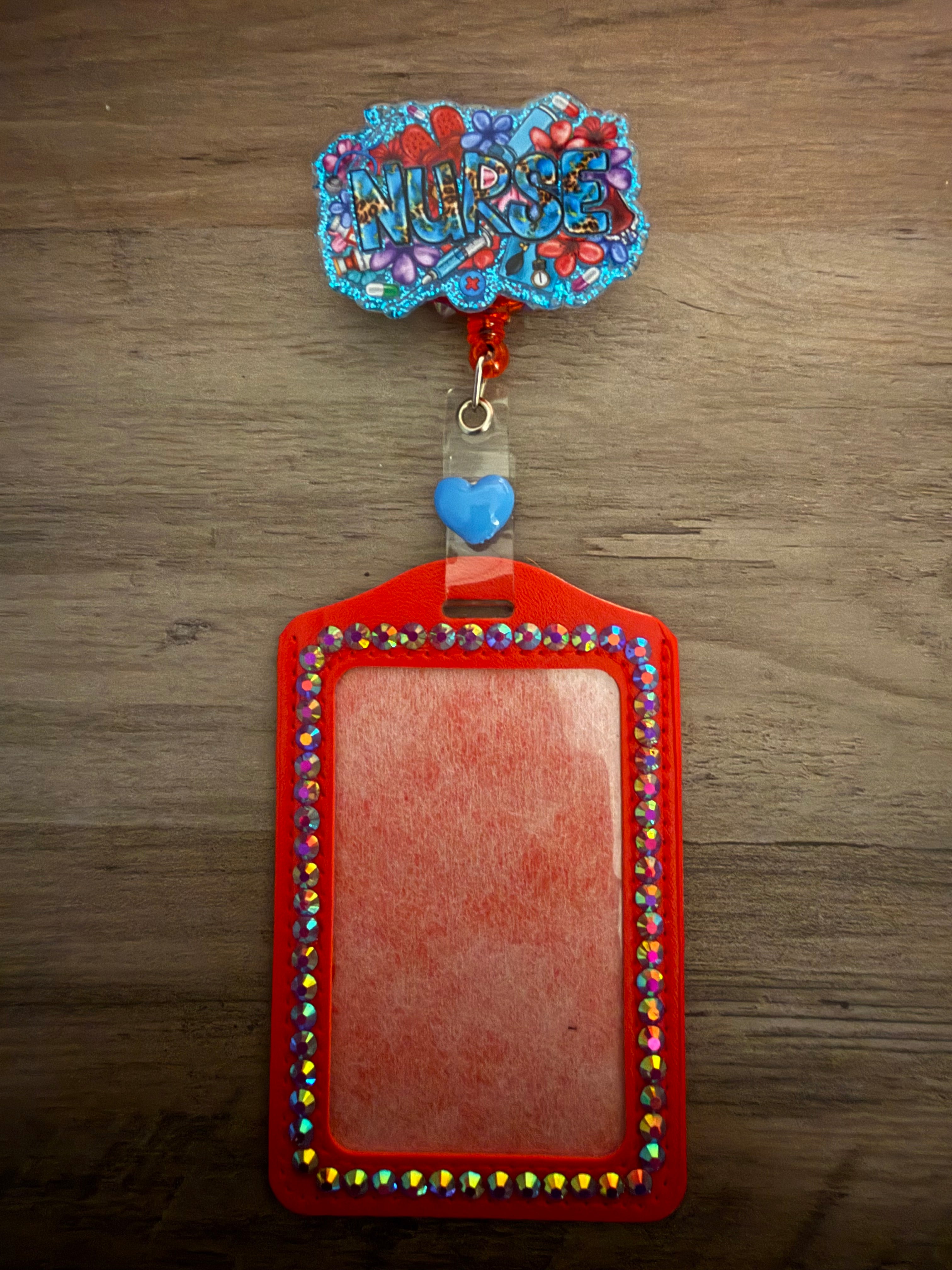 A red ID badge holder bordered with shimmering blue and iridescent rhinestones, attached to a glittery blue retractable reel featuring the word "NURSE" surrounded by colorful medical and floral illustrations.