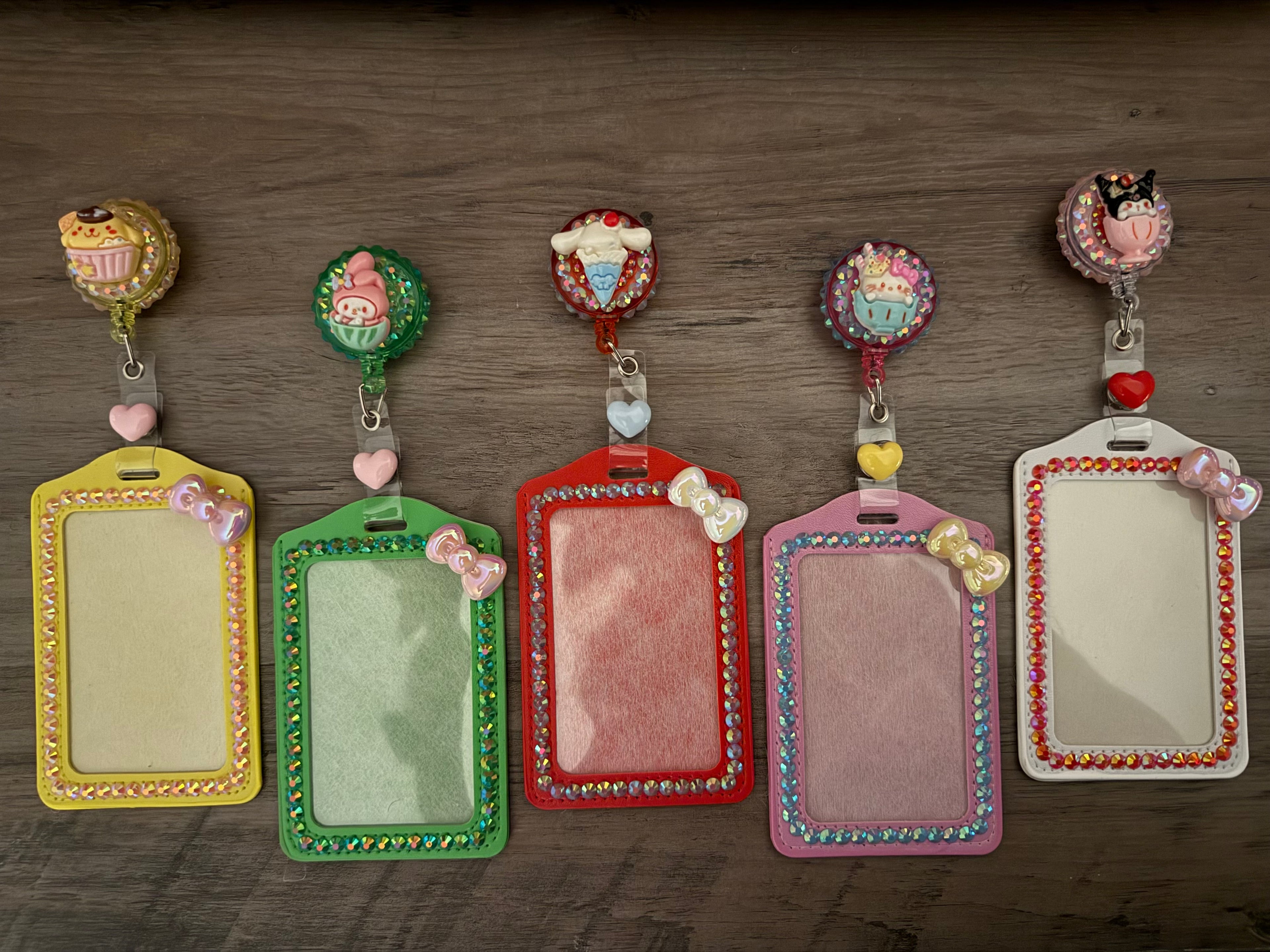 Five colorful, rhinestone-embellished ID card holders with matching retractable badge reels. Each reel features a Sanrio character charm, including Pompompurin, My Melody, Cinnamoroll, Hello Kitty, and Kuromi, set against a shimmering background. The holders are lined with sparkling gems and topped with small decorative bows.