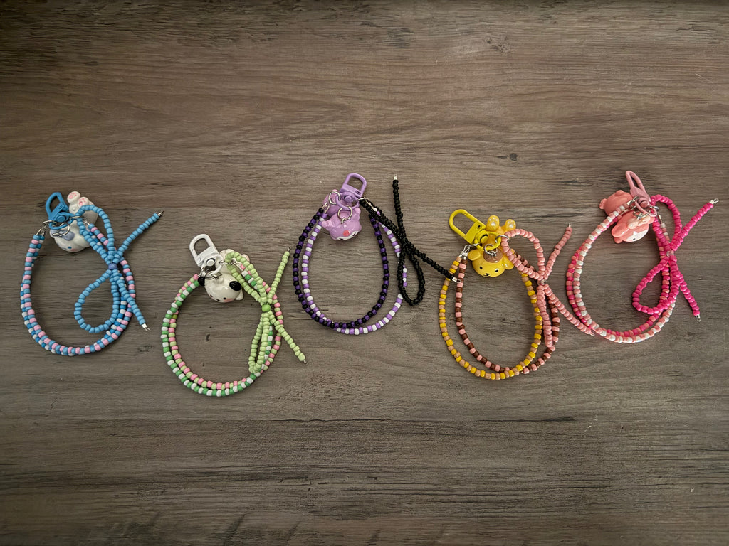 A row of five beaded bag charms on a wooden surface, each featuring a different character charm—Cinnamoroll, Pochacco, Kuromi, Pompompurin, and My Melody—with color-coordinated seed beads and matching lobster clasps.