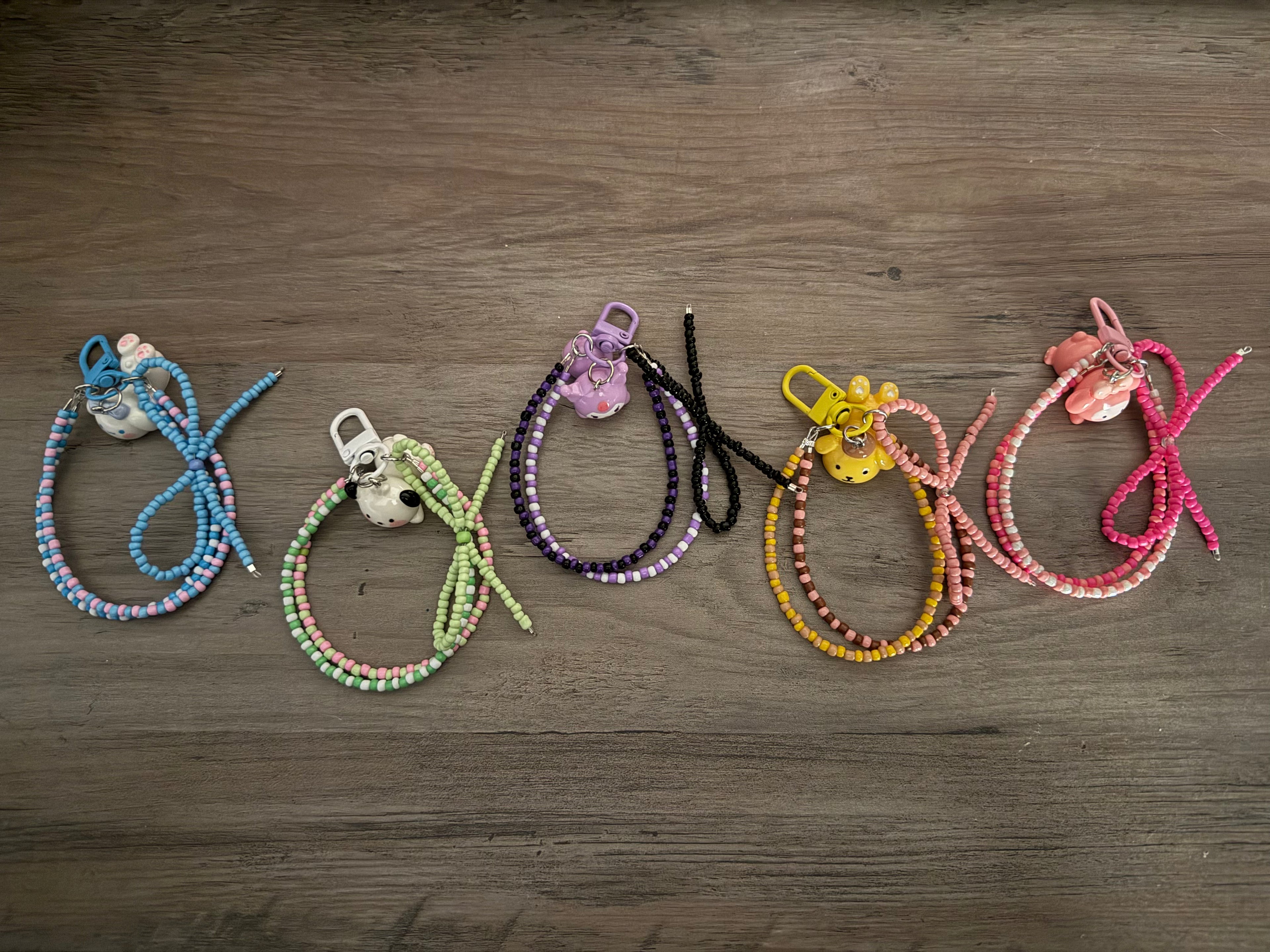 A row of five beaded bag charms on a wooden surface, each featuring a different character charm—Cinnamoroll, Pochacco, Kuromi, Pompompurin, and My Melody—with color-coordinated seed beads and matching lobster clasps.