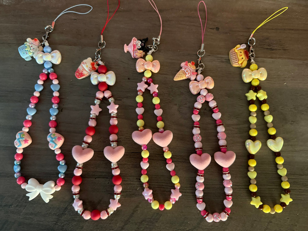 Five handcrafted, beaded phone wristlets in various colors, each featuring a different Sanrio character charm at the top. The wristlets are decorated with a mix of heart-shaped and star-shaped beads, iridescent spacers, and decorative bows. The charms include Hello Kitty, My Melody, Kuromi, and Pompompurin, themed with sweets like ice cream and cupcakes.