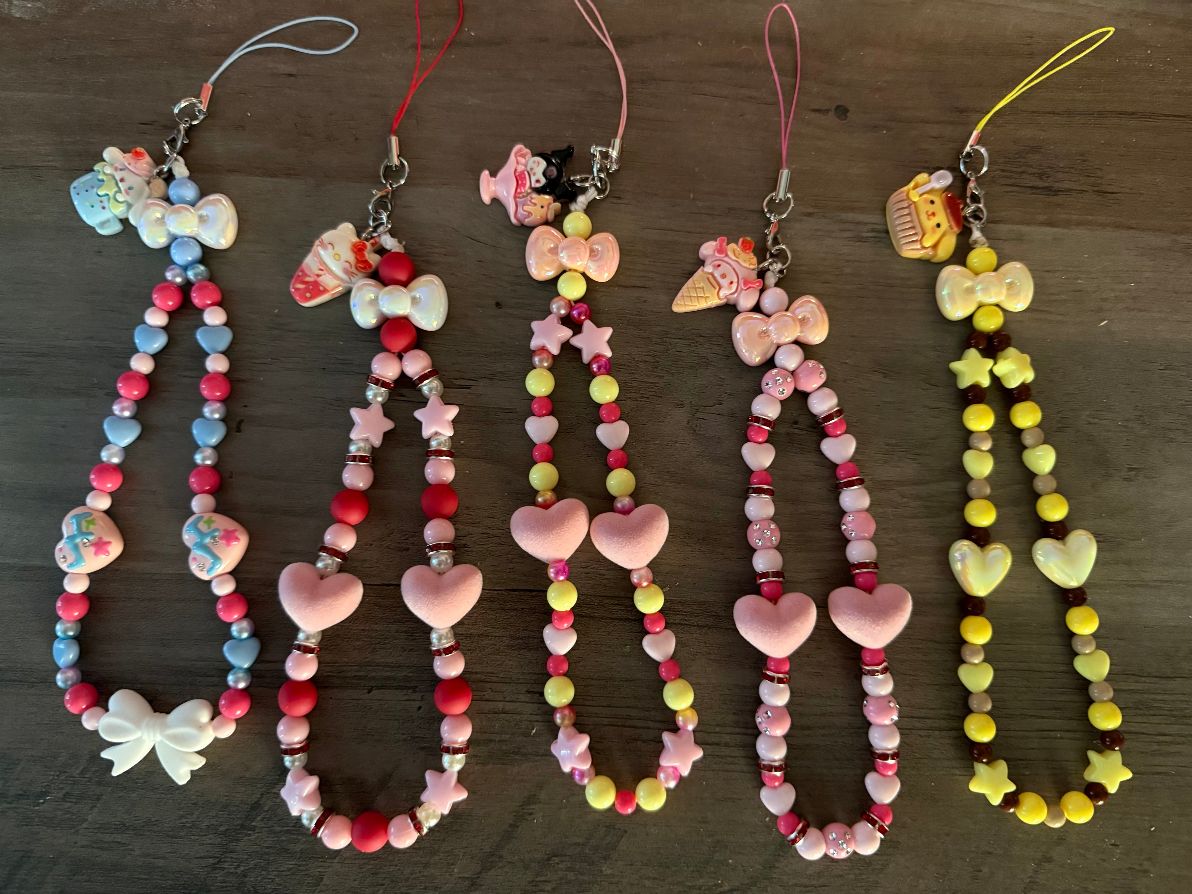Five handcrafted, beaded phone wristlets in various colors, each featuring a different Sanrio character charm at the top. The wristlets are decorated with a mix of heart-shaped and star-shaped beads, iridescent spacers, and decorative bows. The charms include Hello Kitty, My Melody, Kuromi, and Pompompurin, themed with sweets like ice cream and cupcakes.