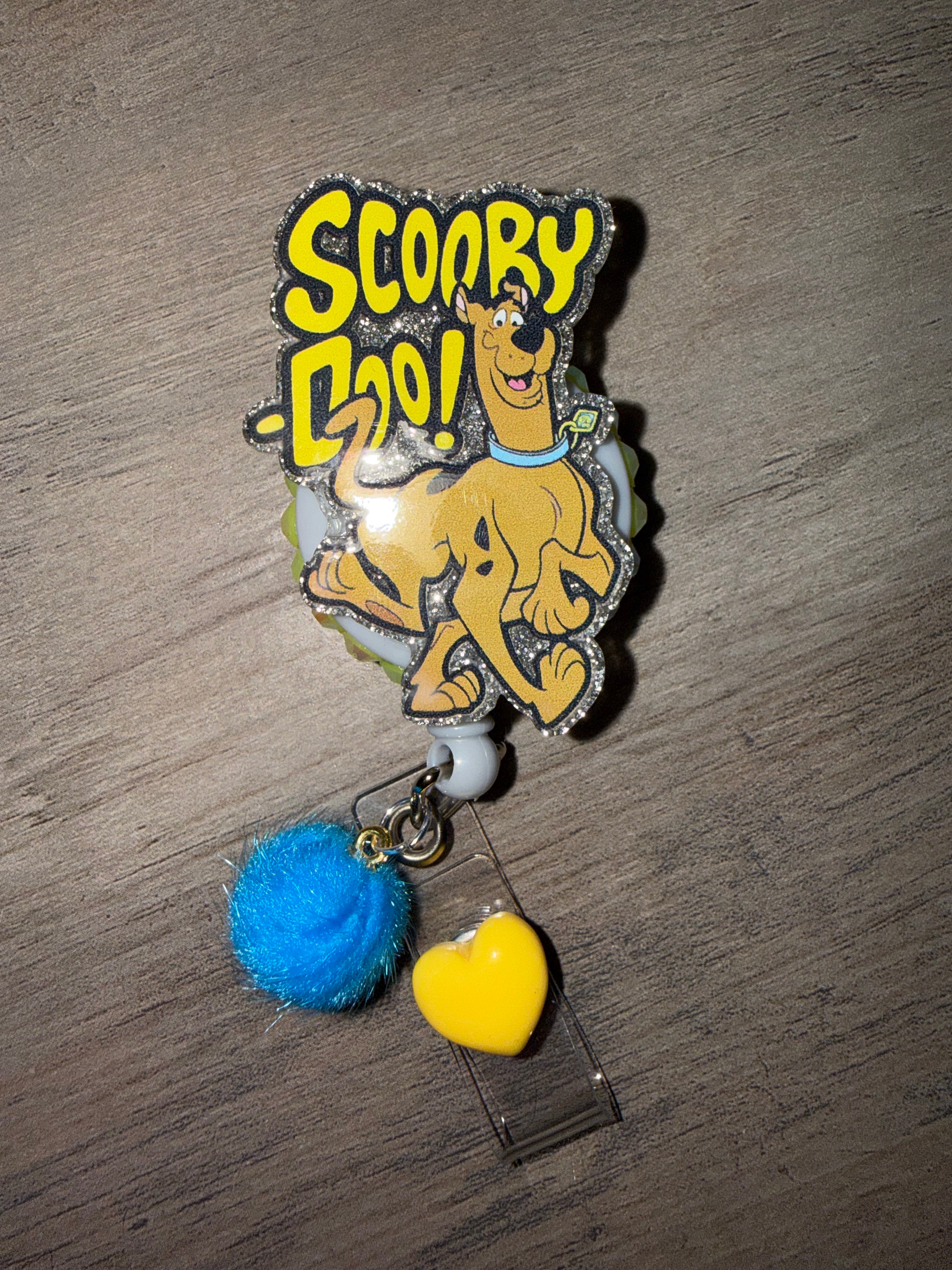 A glittery badge reel featuring a running Scooby-Doo below yellow "Scooby-Doo!" text. The charm is bordered with silver glitter and attached to a white retractable cord, decorated with a small blue pom-pom and a yellow heart charm hanging from the clear strap.