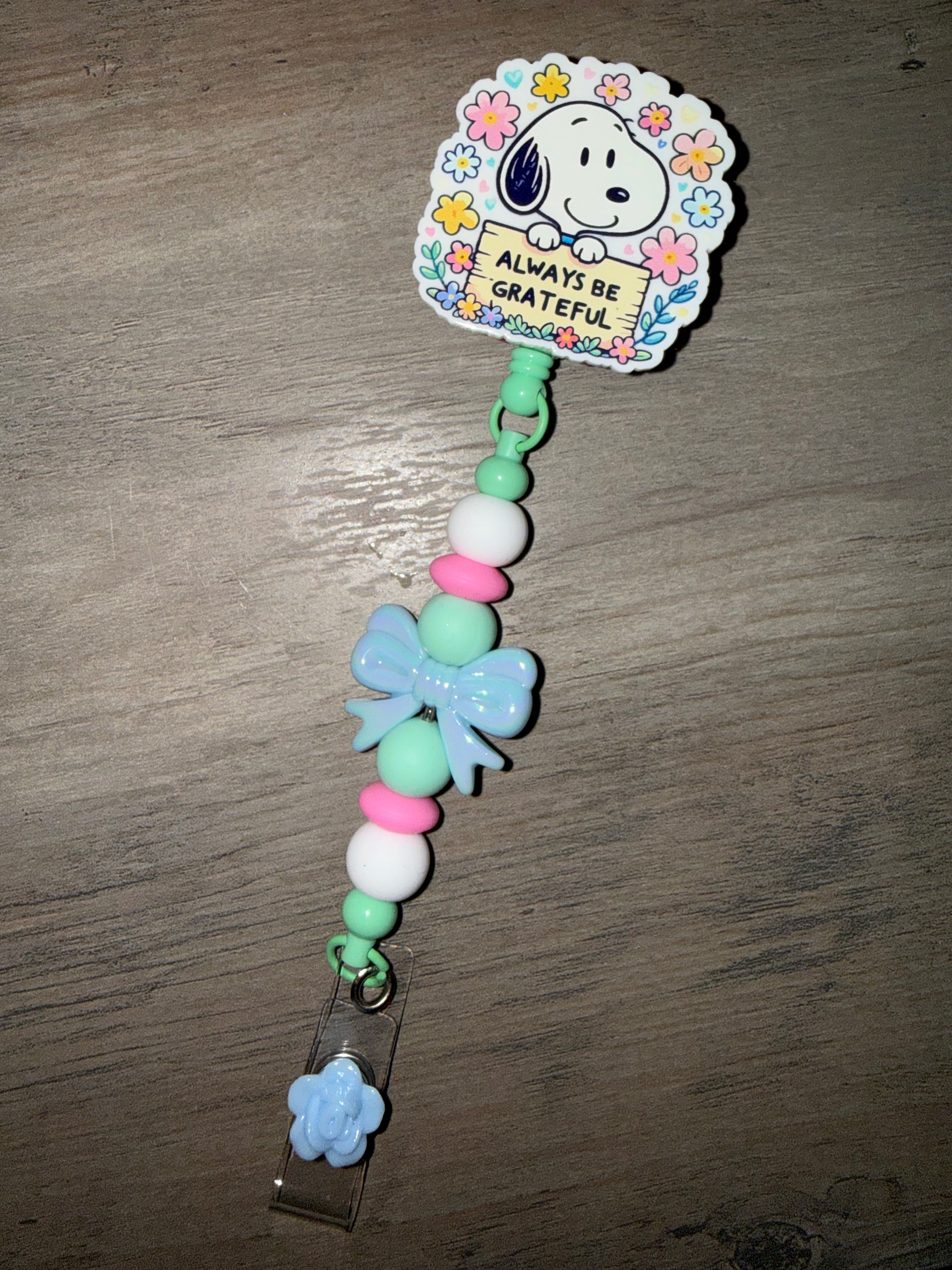 A pastel-colored beaded badge reel featuring Snoopy holding a sign that reads "ALWAYS BE GRATEFUL" surrounded by colorful flowers. The lanyard cord is decorated with mint green, white, and pink beads, a large light blue bow charm, and a small blue flower clip at the bottom.