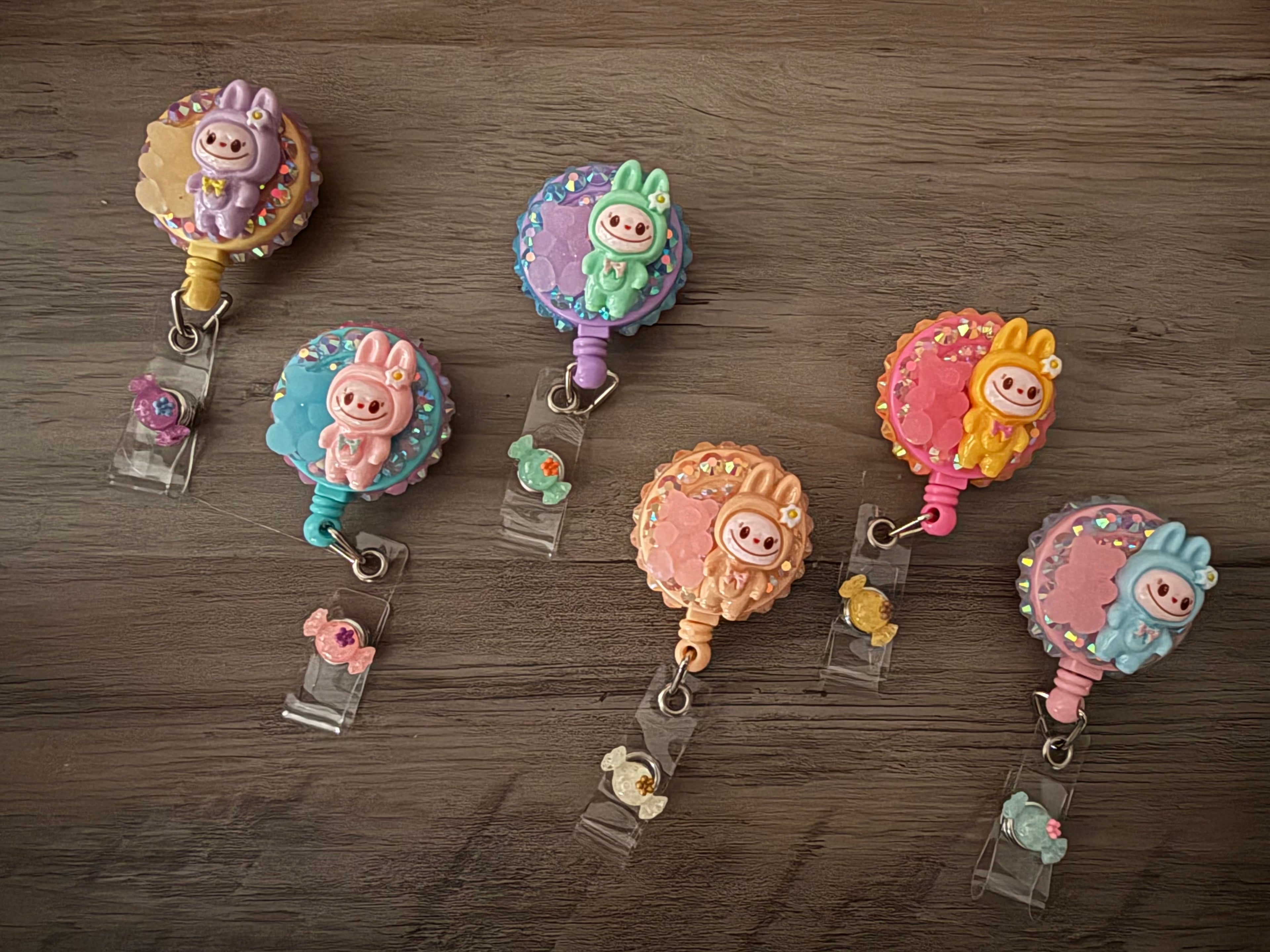 A collection of six colorful, decorative badge reels, each featuring a monster figure in a pastel bunny costume surrounded by glitter and small candy or bear charms.