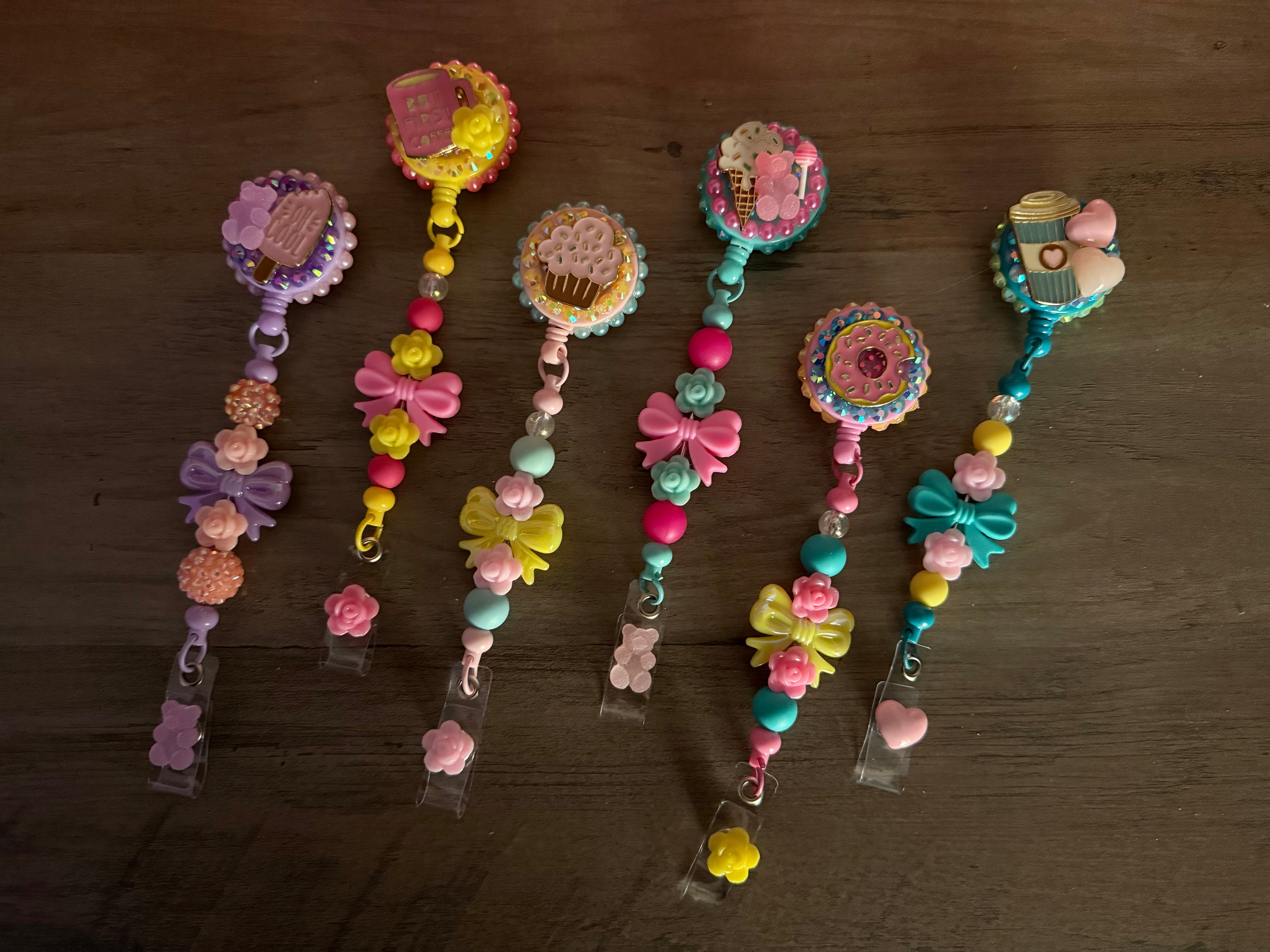 A collection of six colorful, handmade "kawaii" badge reels featuring sweet treat charms like cupcakes, donuts, and coffee, accented with elaborate beaded lanyards, bows, and gummy bear dangles.