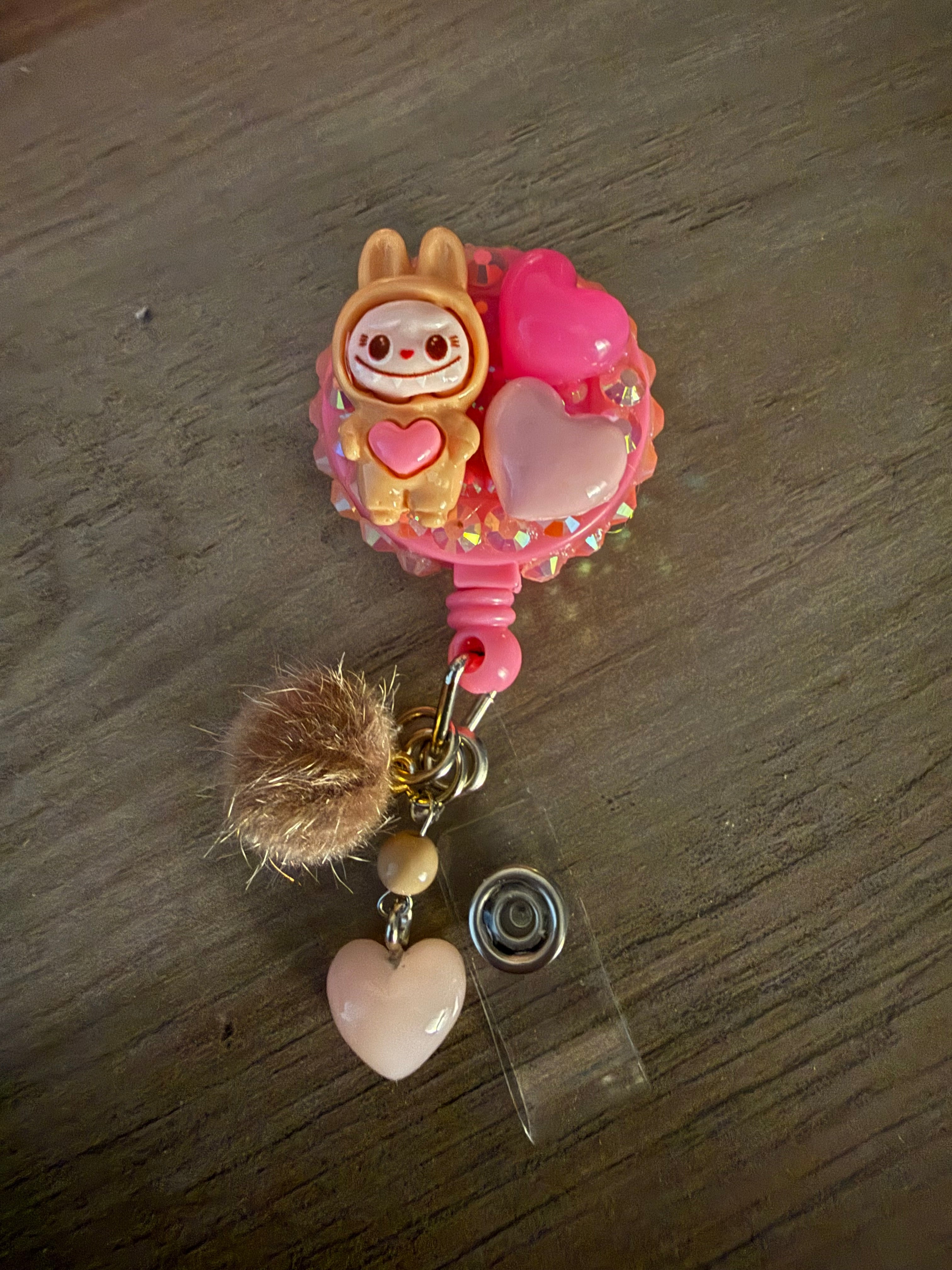 A pink and tan handcrafted decoden badge reel featuring a small tan monster figure, pink resin hearts, and iridescent rhinestones, finished with a tan pom-pom and a light pink heart charm.