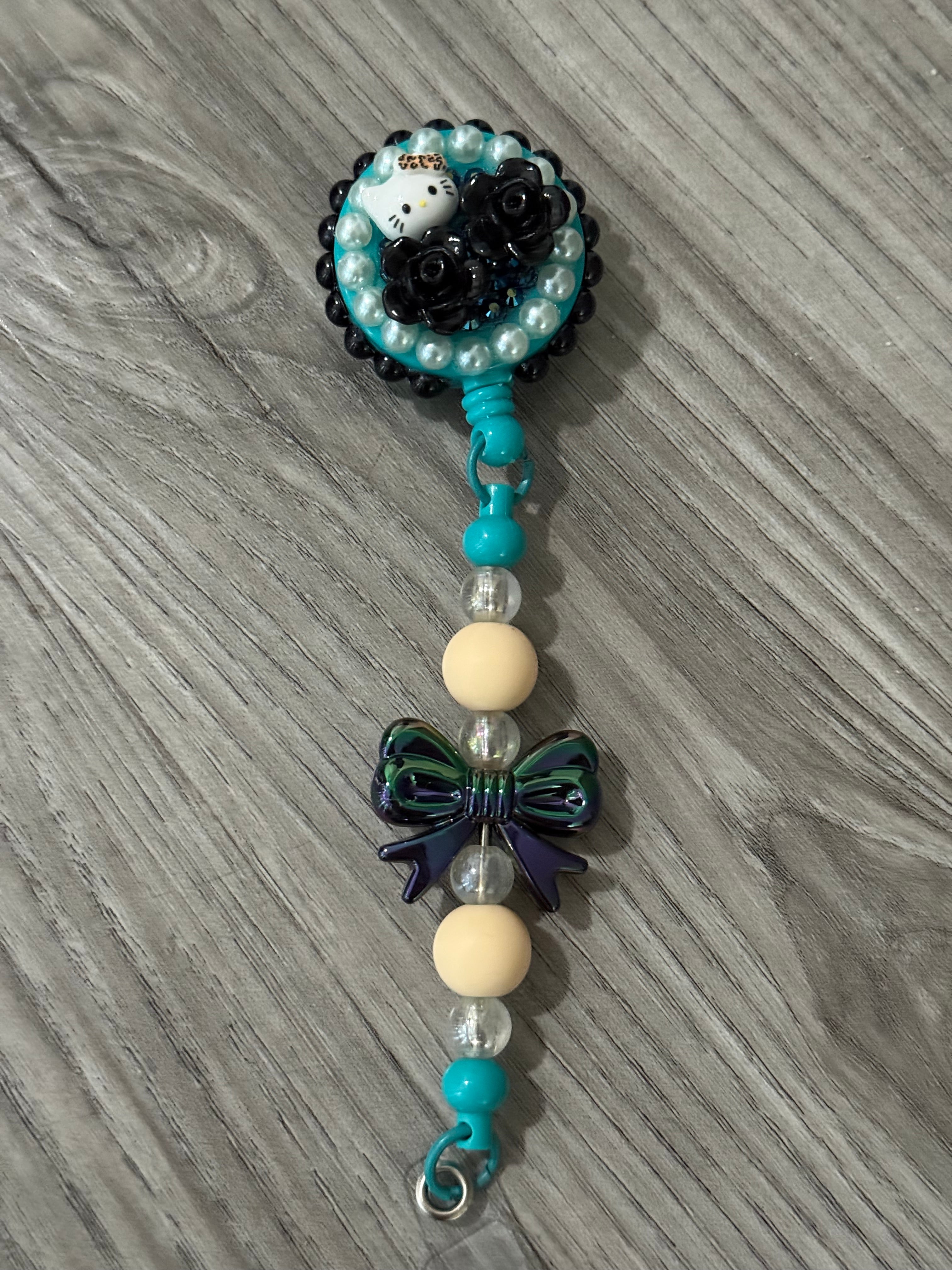 Cute badge reel featuring a playful design with a Hello Kitty inspired character, black roses, and turquoise accents, complemented by a decorative bow and colorful beads.