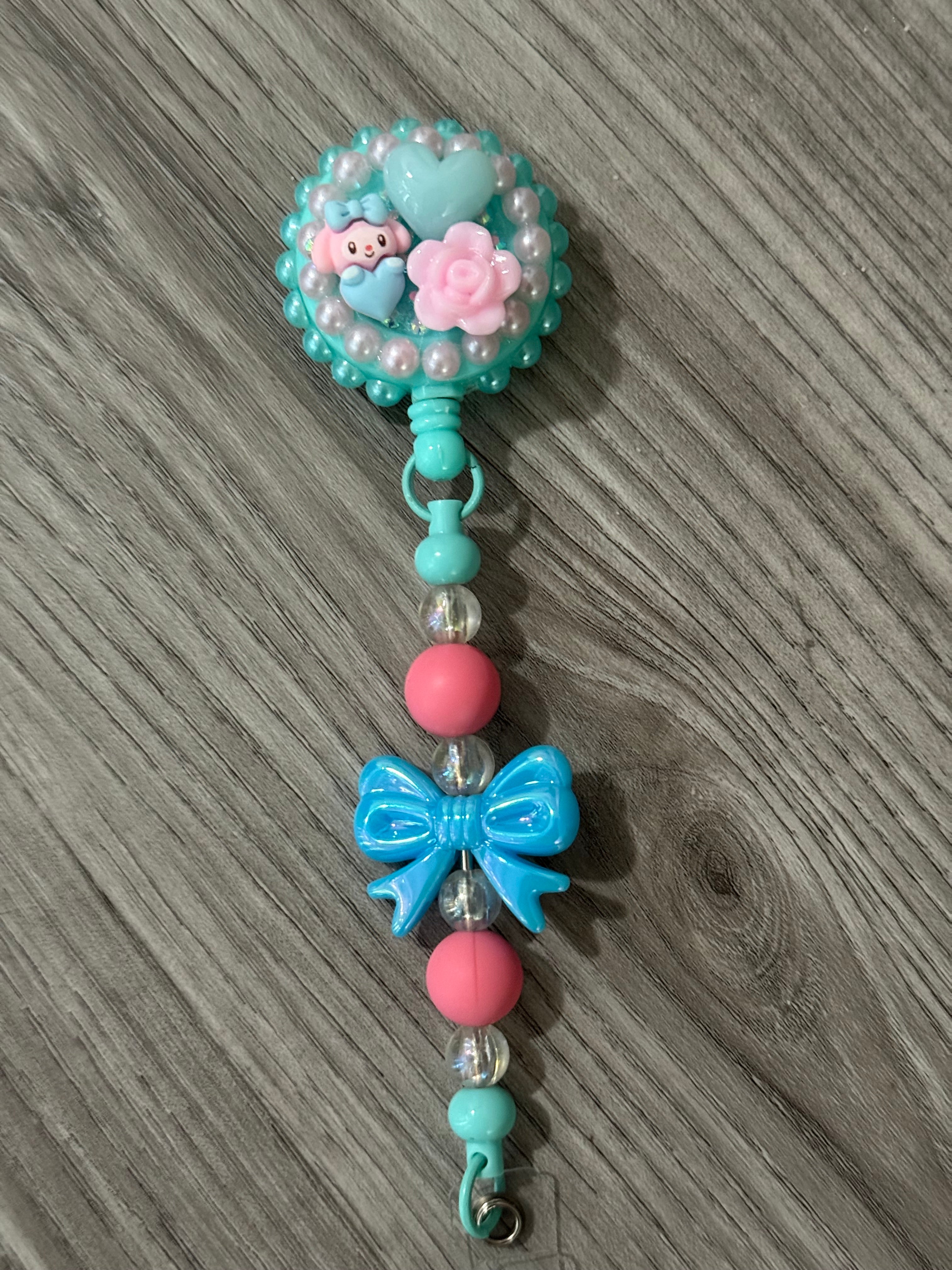 Cute badge reel featuring a playful design with a turquoise top adorned with a my melody inspired character, pink flowers, and hearts, complemented by colorful beads and a blue bow on the strap. 