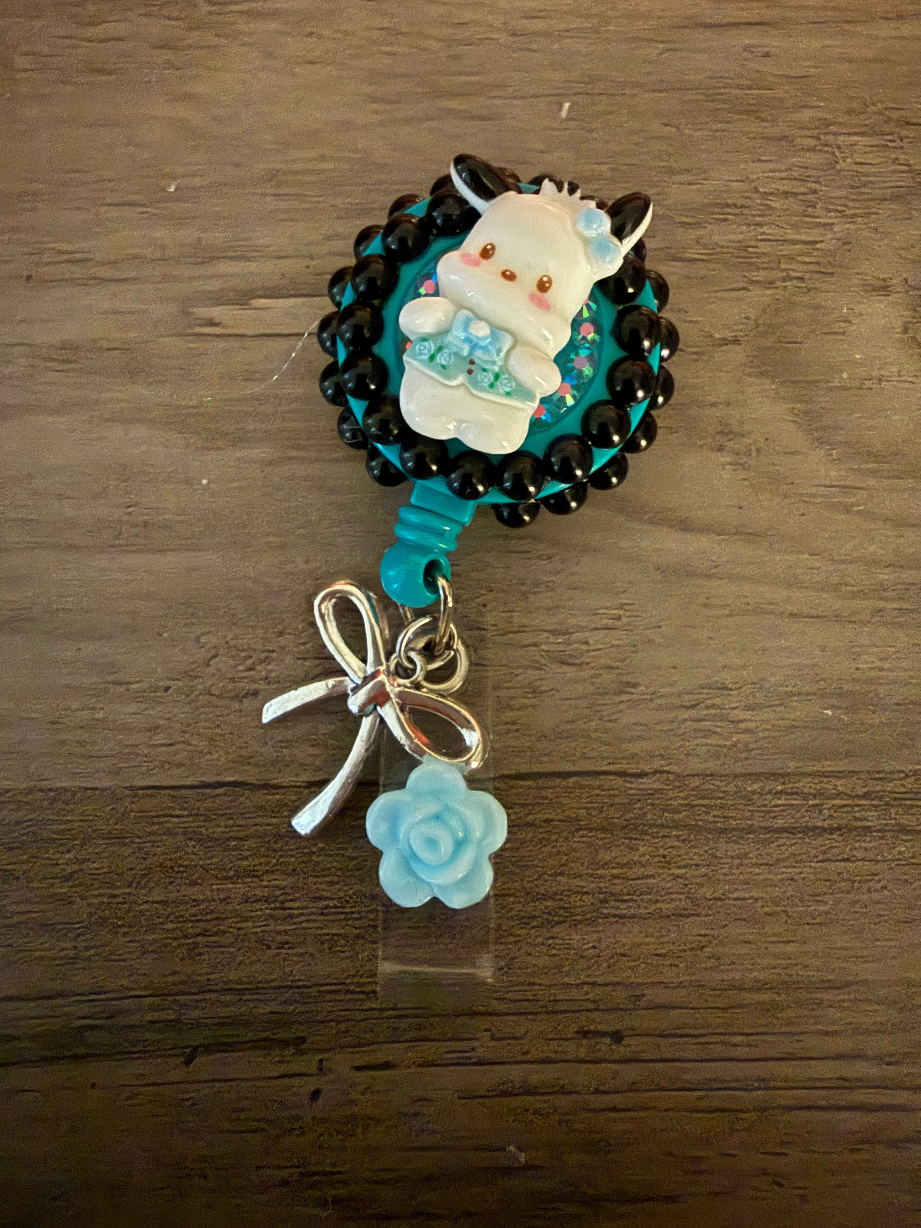 Colorful badge reel featuring a cute pochacco inspired charm with a teal background, decorative beads, a silver bow, and a light blue rose accent.