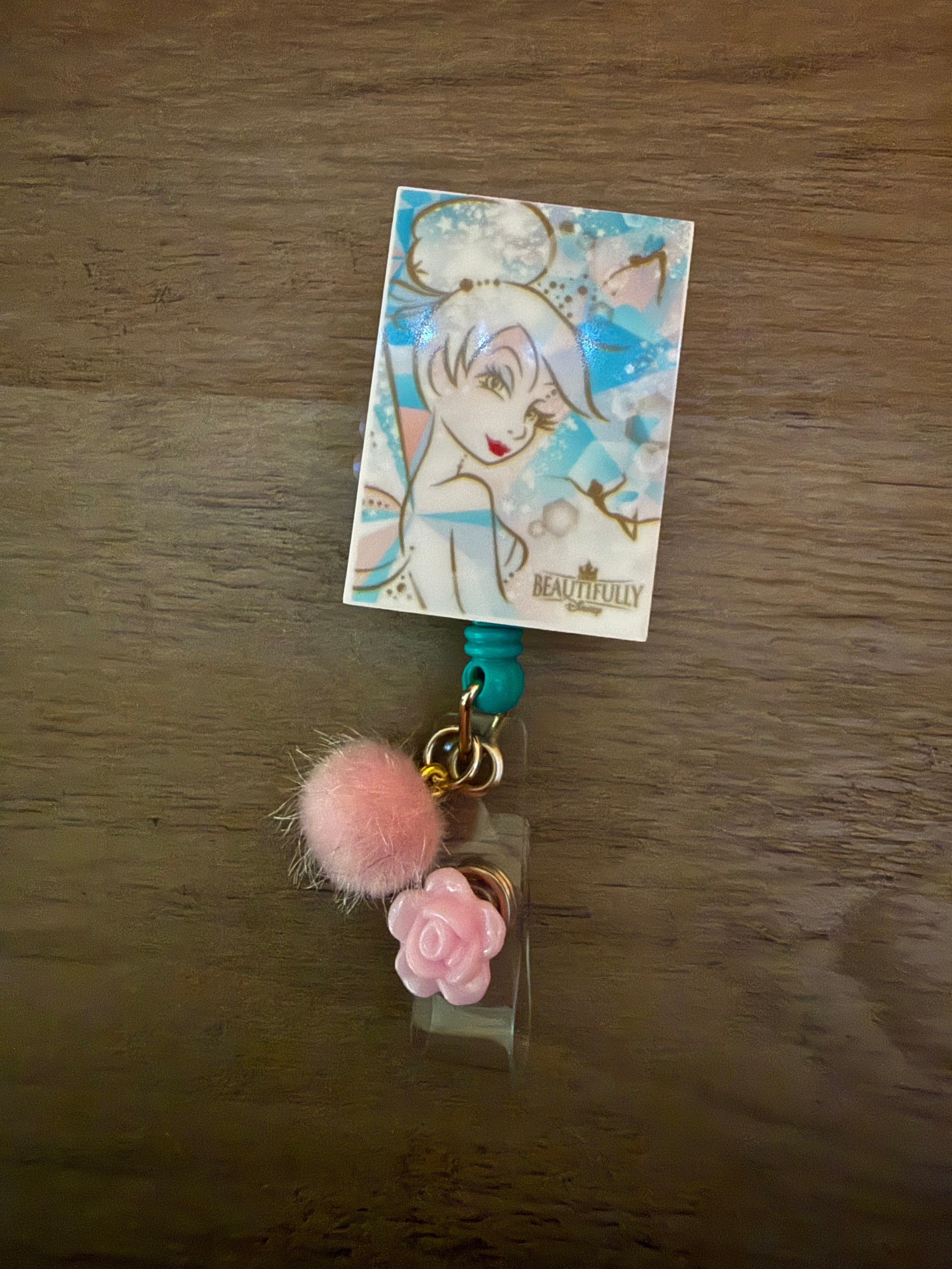 A handcrafted rectangular badge reel featuring a vintage-style Tinker Bell illustration on a blue-and-white background. The reel includes a soft pink fuzzy pom-pom and a small pink rose charm.