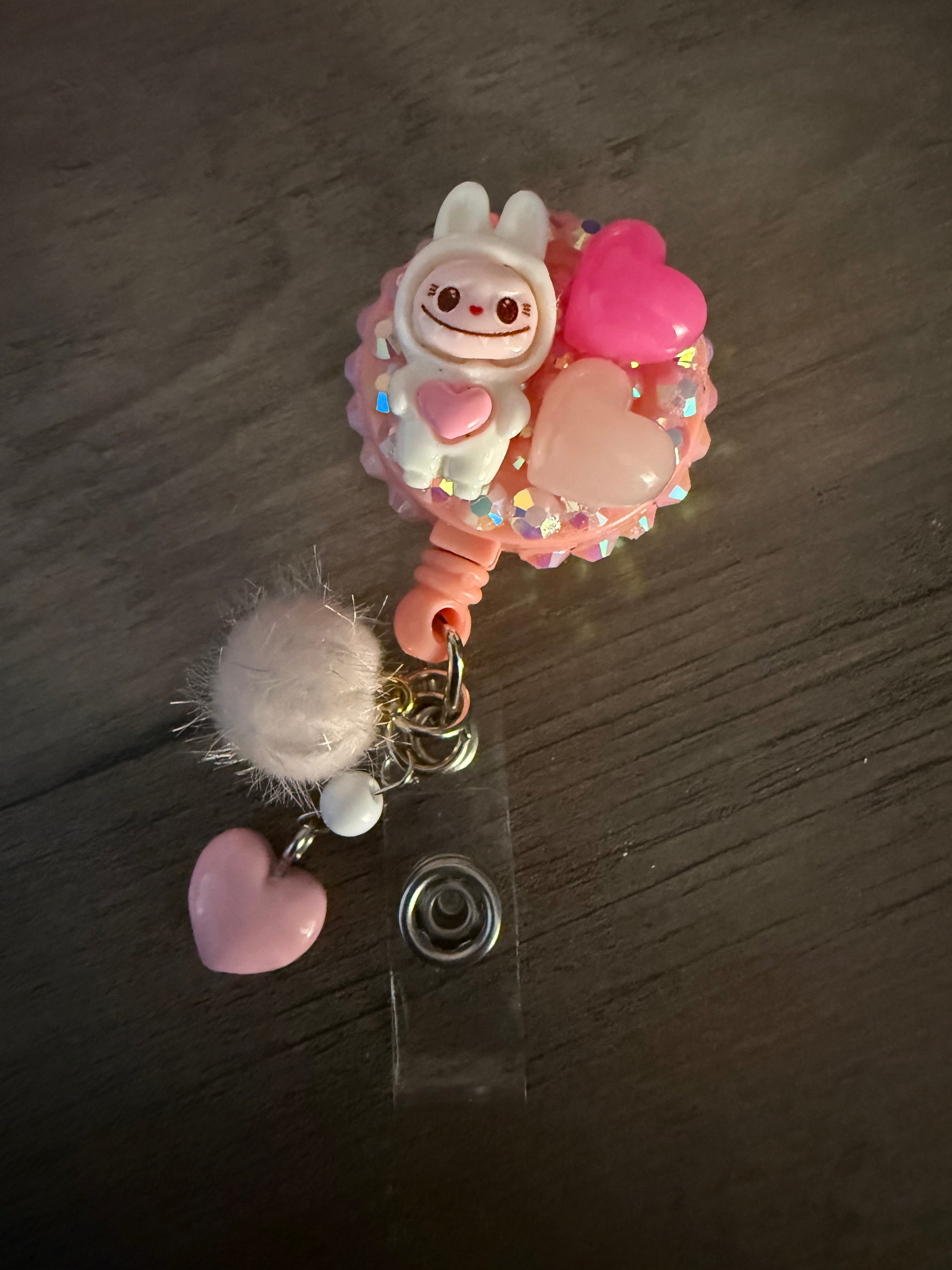 A handcrafted, pink decoden-style retractable badge reel featuring a small white monster holding a heart, surrounded by pink heart charms and iridescent rhinestones. A fluffy pink pom-pom and heart charm hang from the bottom loop.