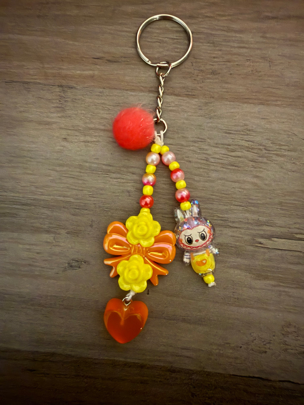 A handcrafted yellow and orange keychain featuring two beaded strands. It includes a fuzzy red pom-pom, a cartoon monster character in yellow overalls, an orange bow decorated with yellow roses, and a translucent orange heart charm.