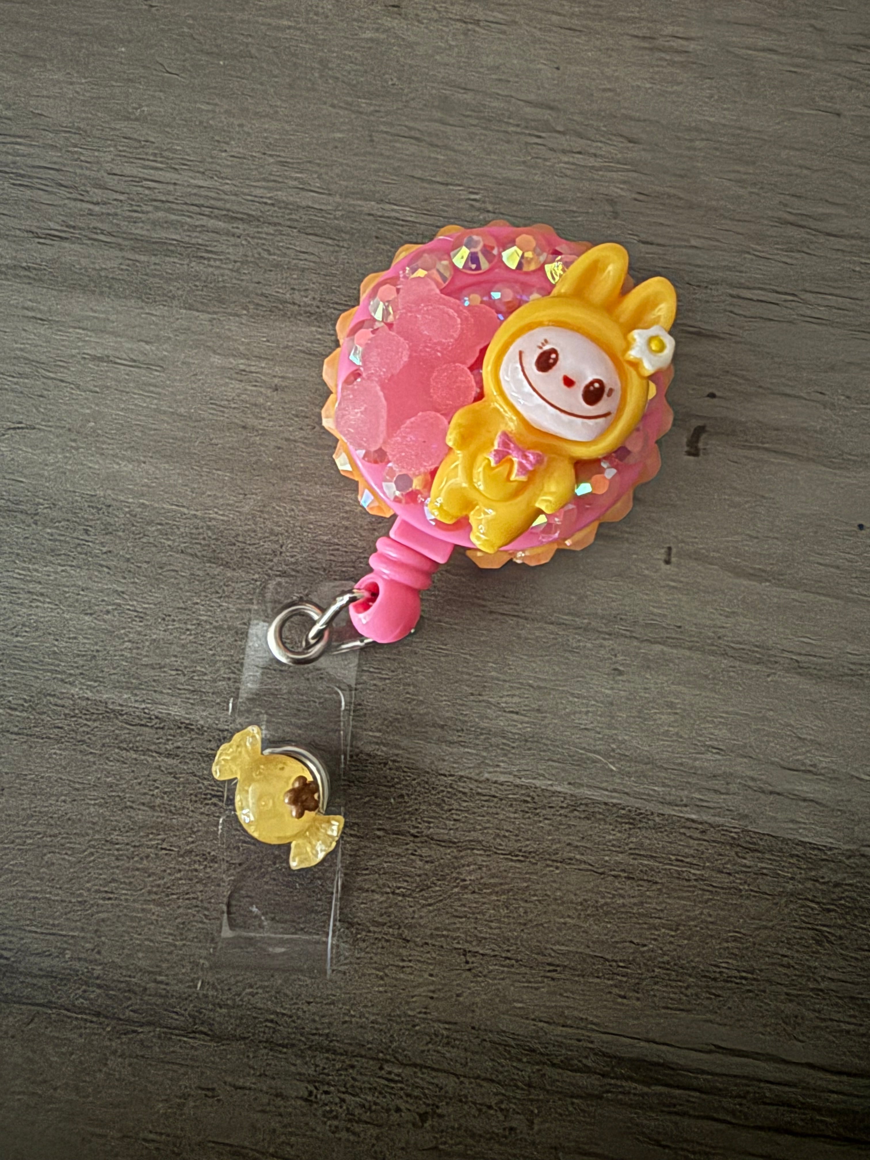 Handcrafted yellow monster in a hooded outfit on a bright pink retractable badge reel, adorned with iridescent rhinestones, a pink gummy bear charm, and a yellow candy charm on the strap.
