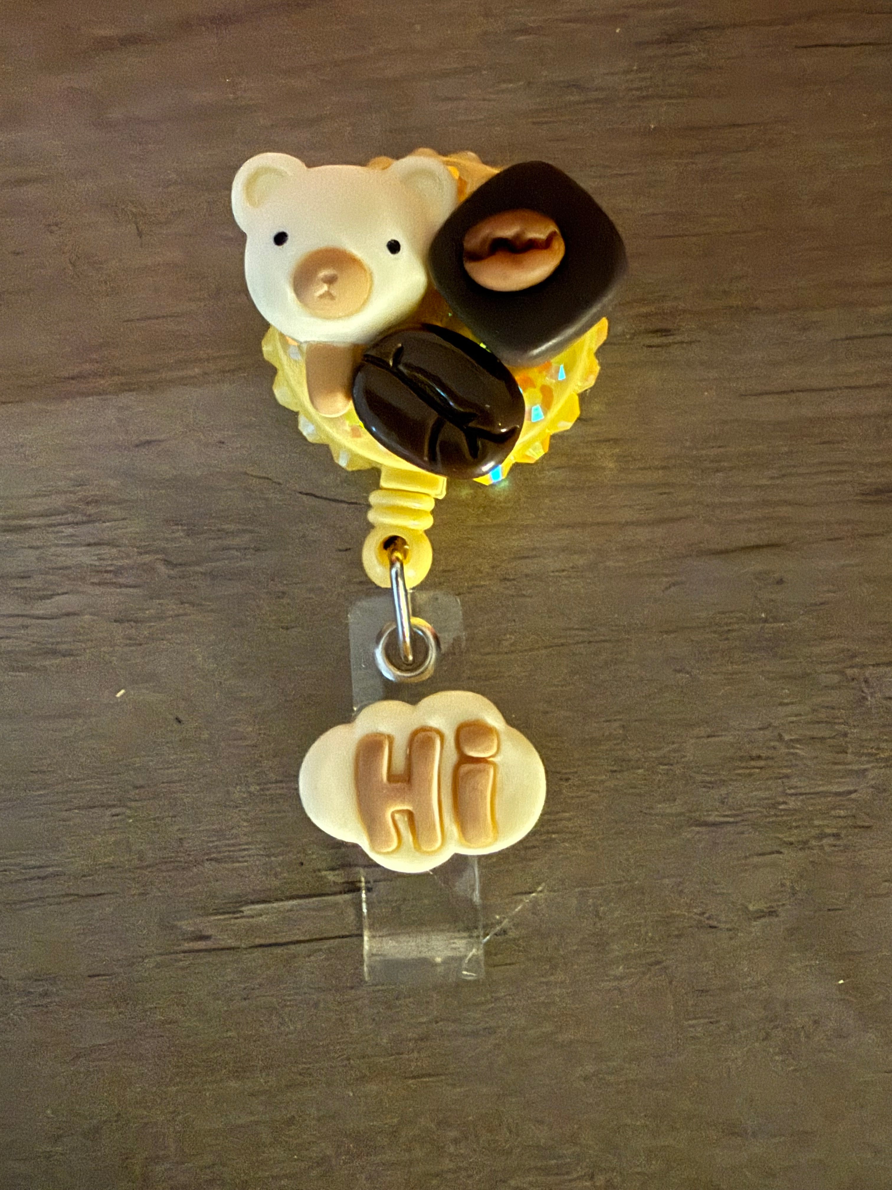 A retractable badge reel featuring a decoden-style yellow glitter base decorated with a white bear charm and coffee bean accents, with a white cloud charm that says "HI" hanging from the clip.