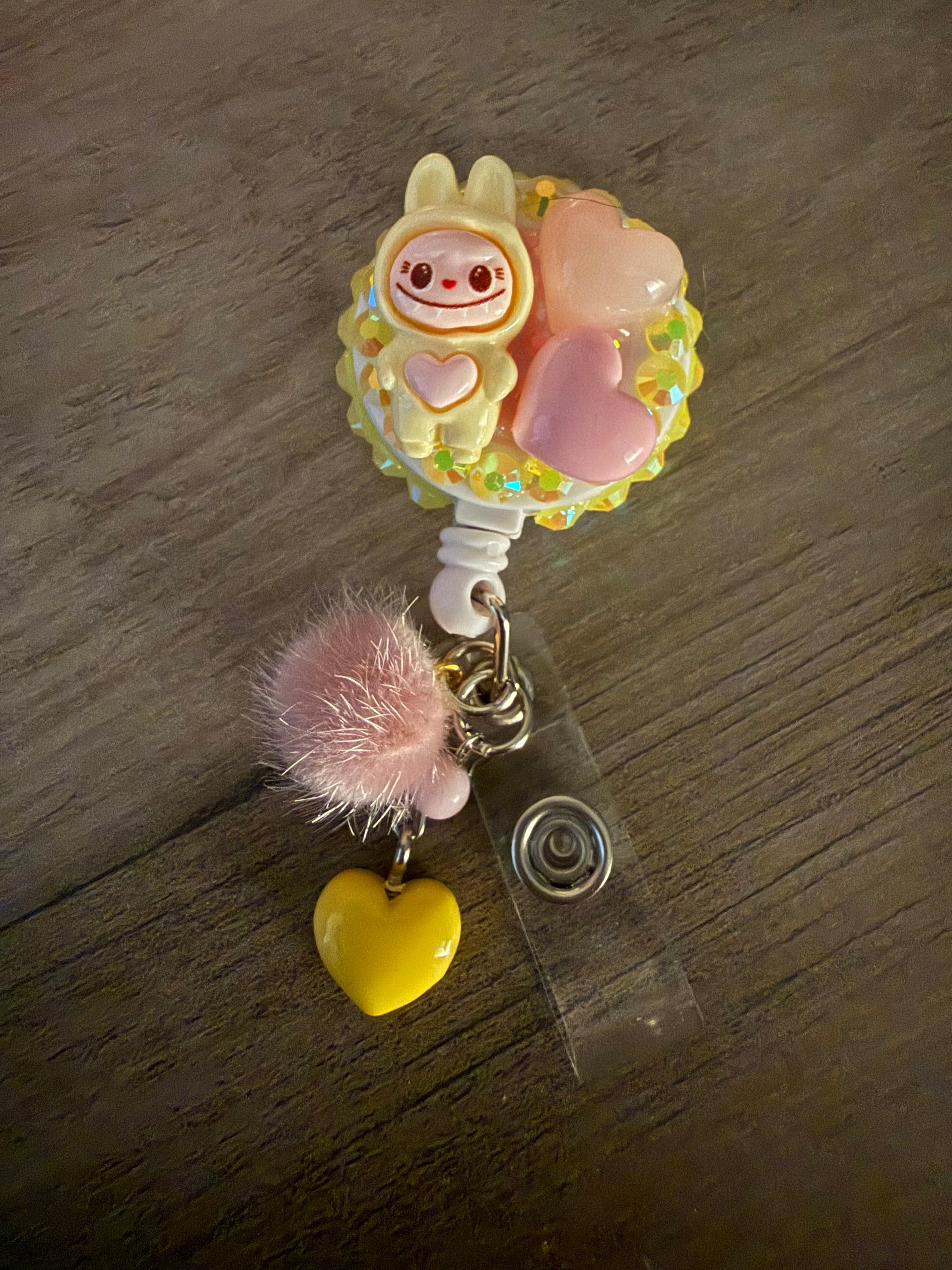 A handcrafted decoden badge reel featuring a yellow rabbit-costume monster, pink hearts, and sparkling rhinestones. Attached is a pink pom-pom and a yellow heart charm.