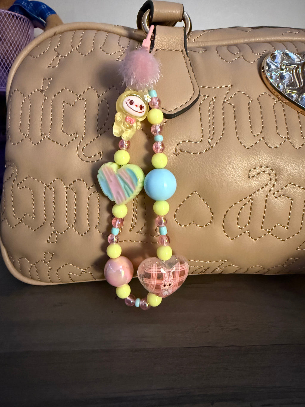 A pastel beaded bag charm with yellow, pink, and blue accents attached to a tan quilted handbag with stitched lettering and a crystal heart detail.