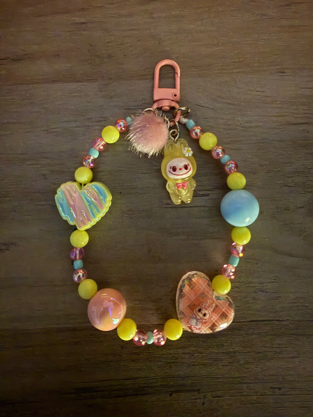 A pastel beaded bag charm featuring yellow, pink, and blue beads, a pink pom-pom, a heart-shaped pendant, and a character charm, attached to a pink lobster clasp on a dark wood background.