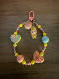 A pastel beaded bag charm featuring yellow, pink, and blue beads, a pink pom-pom, a heart-shaped pendant, and a character charm, attached to a pink lobster clasp on a dark wood background.