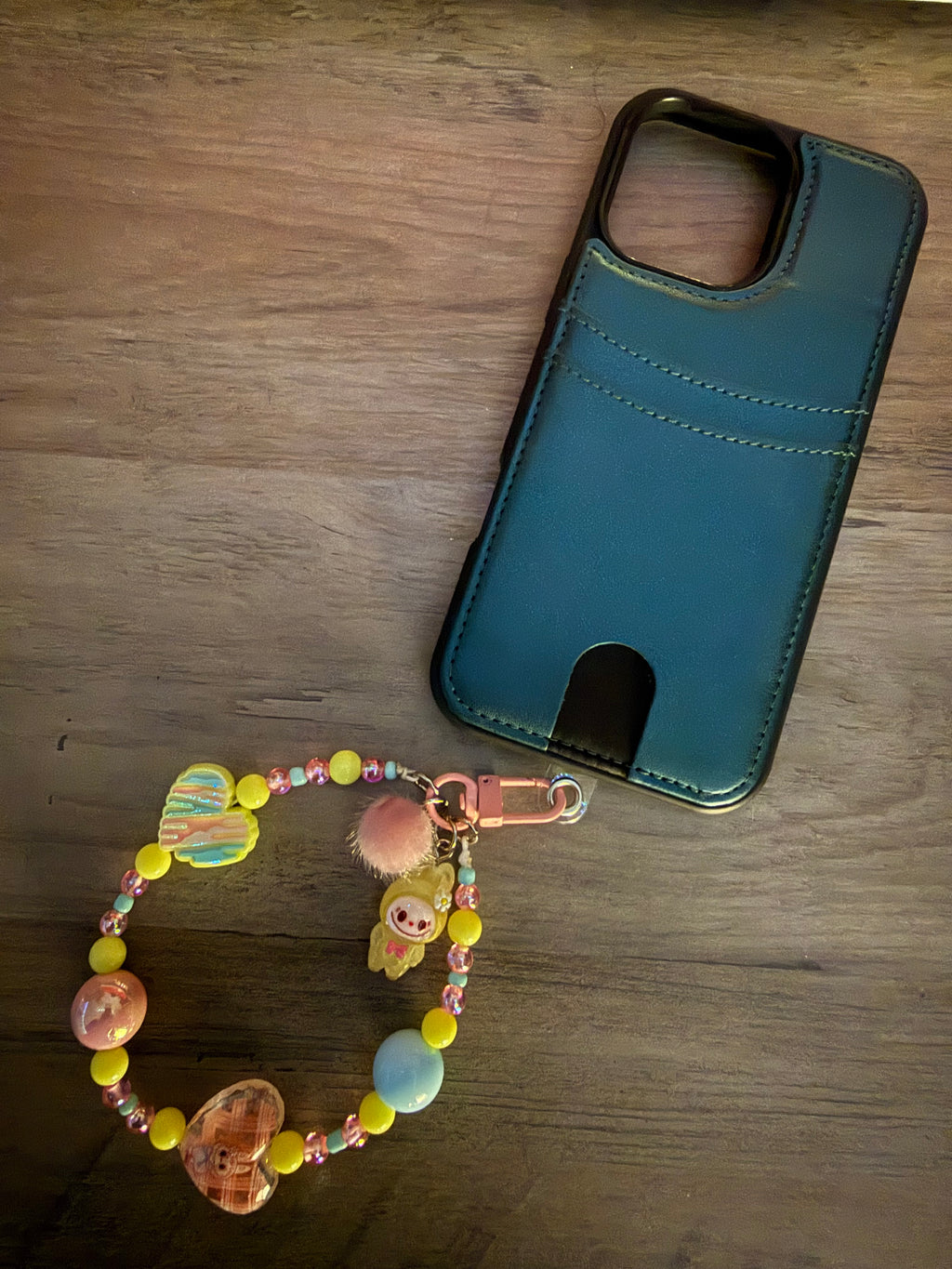 A pastel beaded phone charm with yellow and pink accents attached to a dark leather-style cardholder phone case.