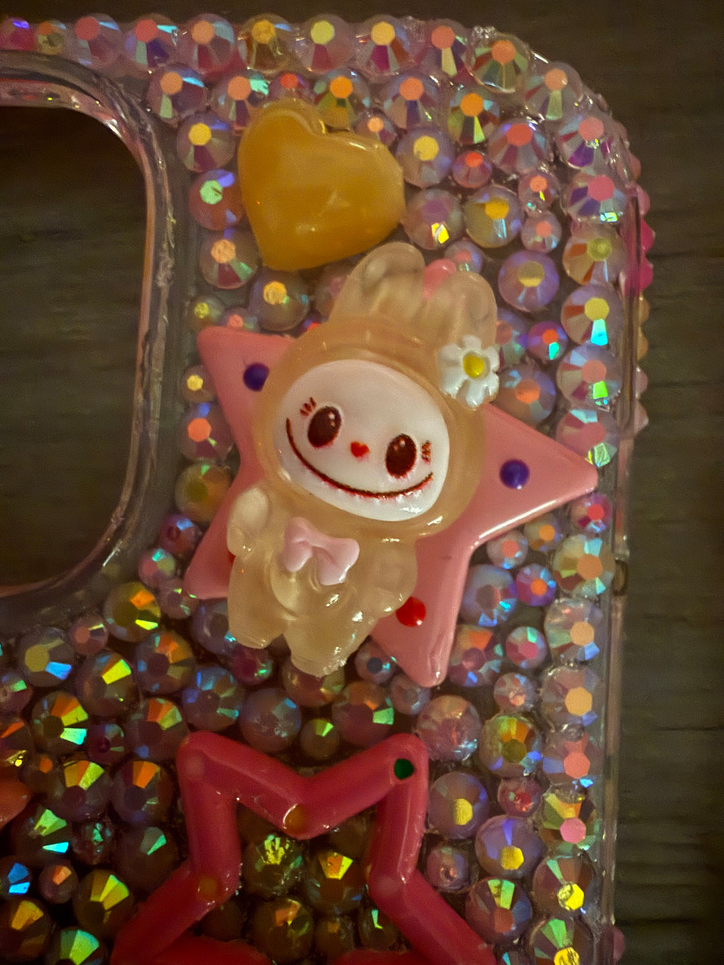 Close-up of a decoden phone case featuring iridescent rhinestones, a pink star cabochon, and a yellow monster character charm with a small flower and a wide smile.
