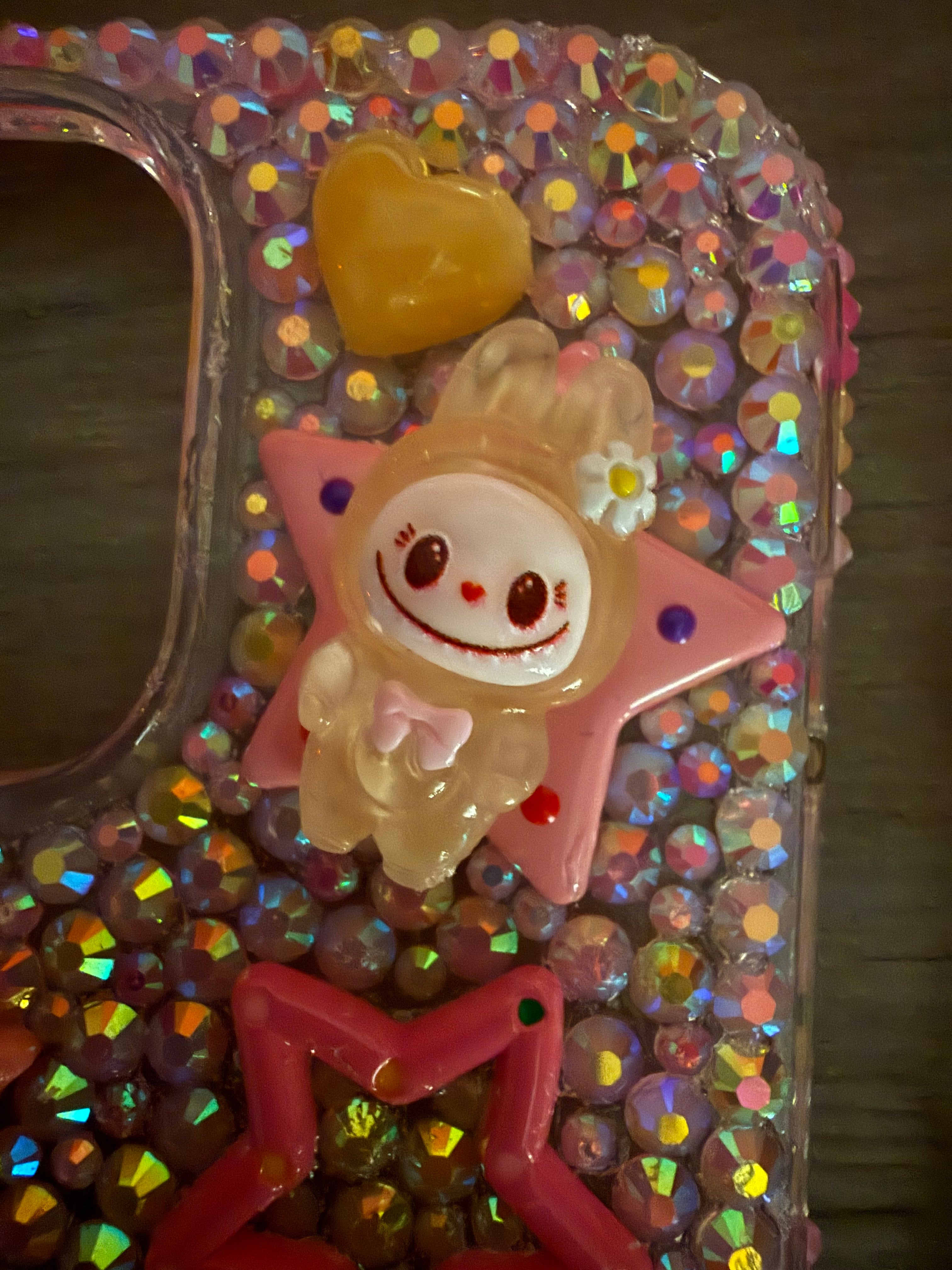 Close-up of a decoden phone case featuring iridescent rhinestones, a pink star cabochon, and a yellow monster character charm with a small flower and a wide smile.