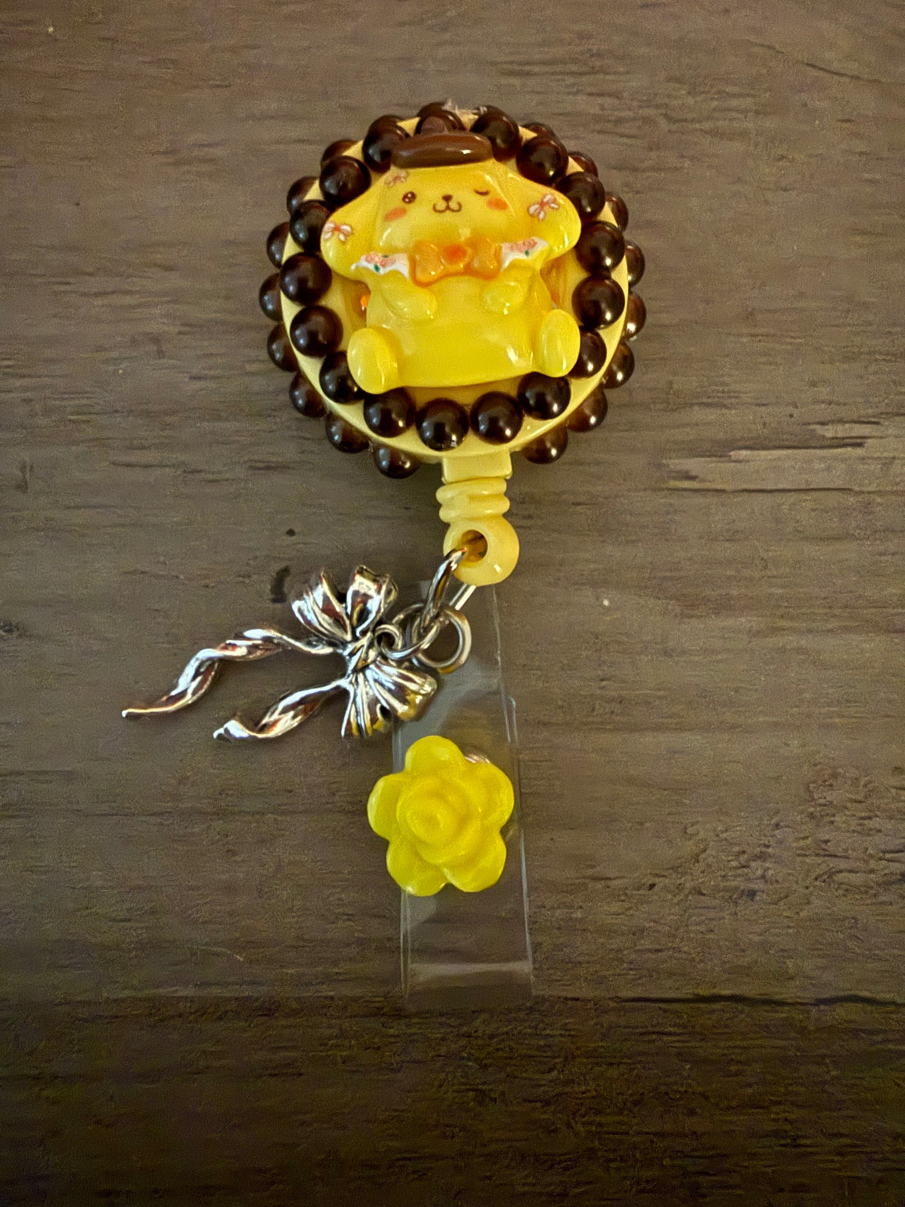 Colorful badge reel featuring a cute yellow pompompurin inspired charm with a brown beaded frame, a silver bow charm, and a small yellow rose accent.