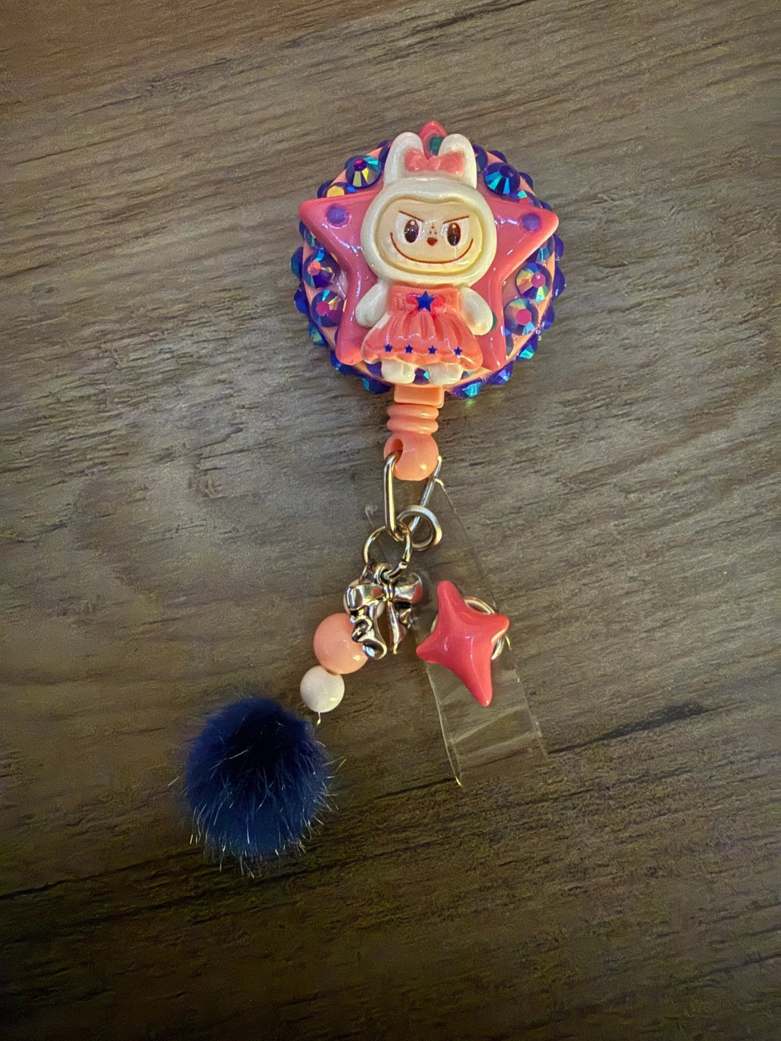 Colorful badge reel featuring a cute character, a star, and playful charms, including a pink pom-pom and beads.