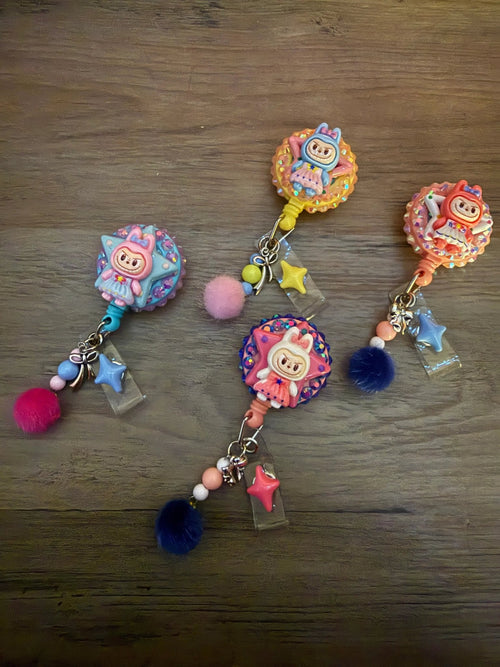 Four colorful badge reels featuring cute character designs. Adorned with playful charms, including pom-poms and stars.
