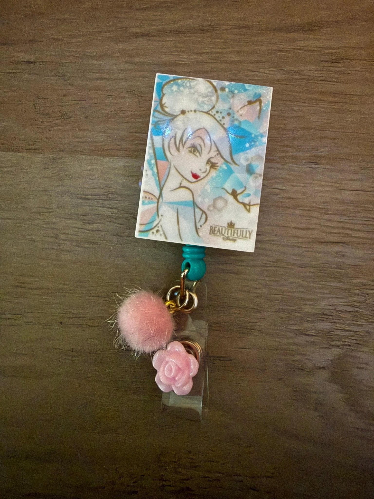 Badge reel featuring a whimsical character illustration, a pink pom-pom, and a small pink rose charm.