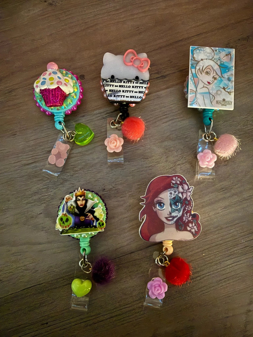 Five colorful badge reels featuring a cupcake, Hello Kitty, a fairy, a villain, and a mermaid, cute charms and pom-poms.