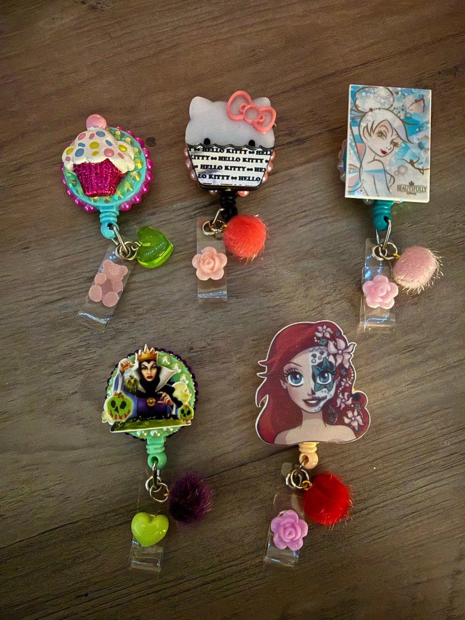 Five colorful badge reels featuring a cupcake, Hello Kitty, a fairy, a villain, and a mermaid, cute charms and pom-poms.