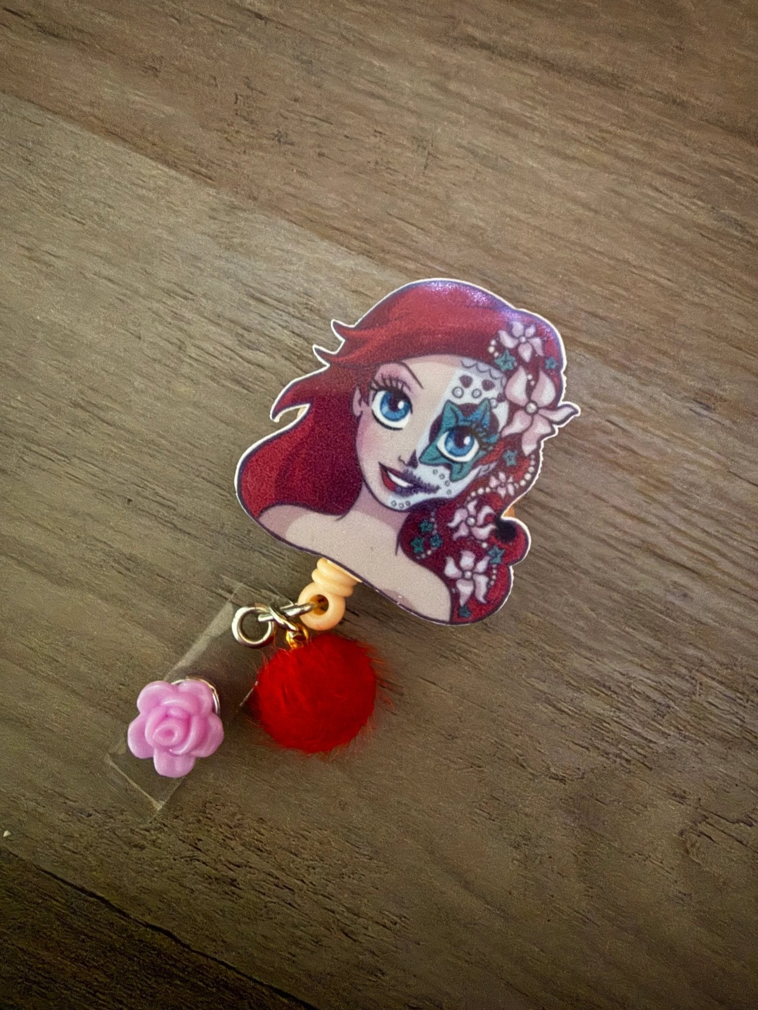 Badge reel featuring a character with red hair, half skull design, pink rose charm, and red pom-pom.