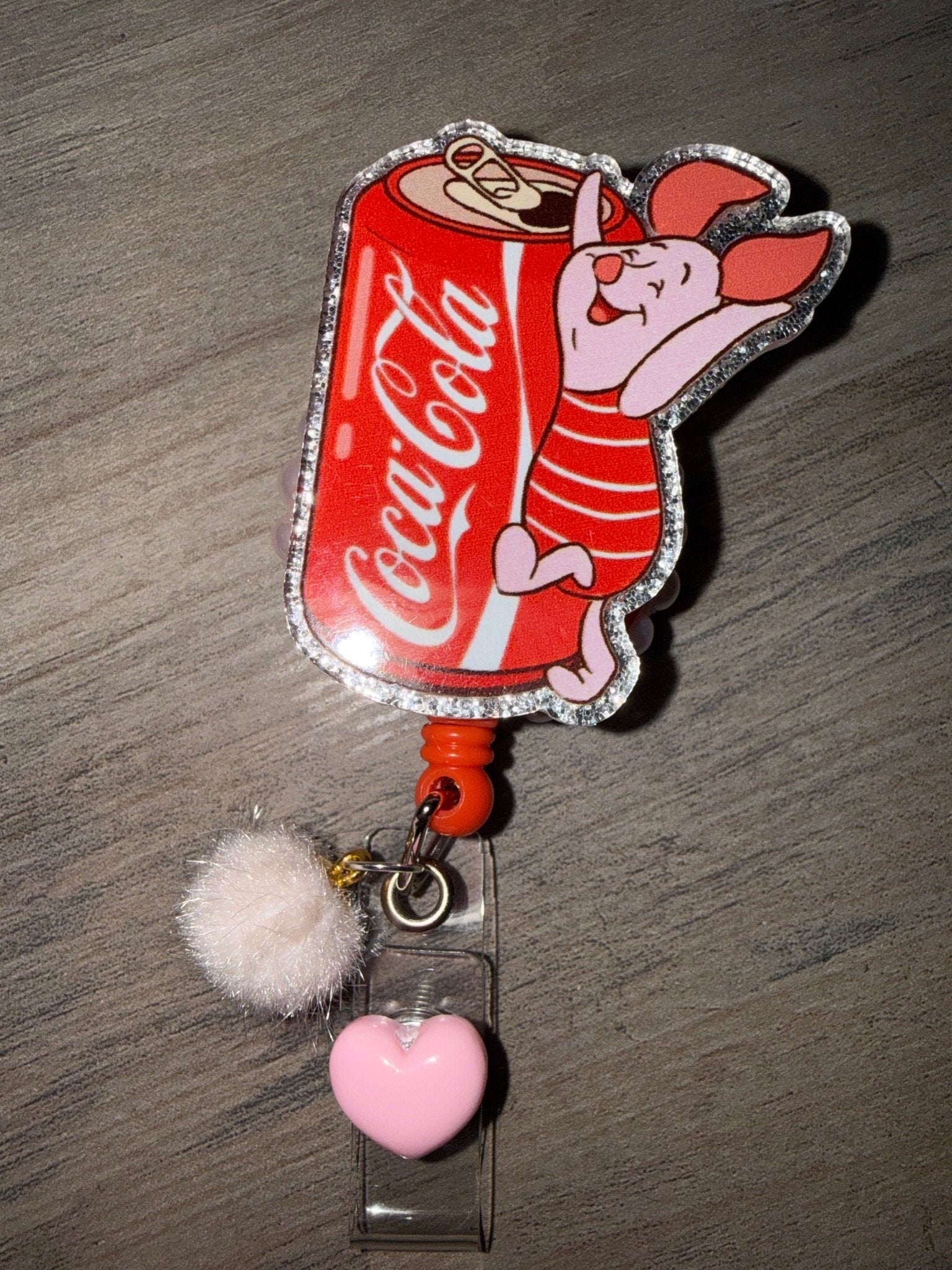 Handmade Piglet and Coca-Cola retractable badge reel with a white fuzzy pom-pom and a pink heart charm.