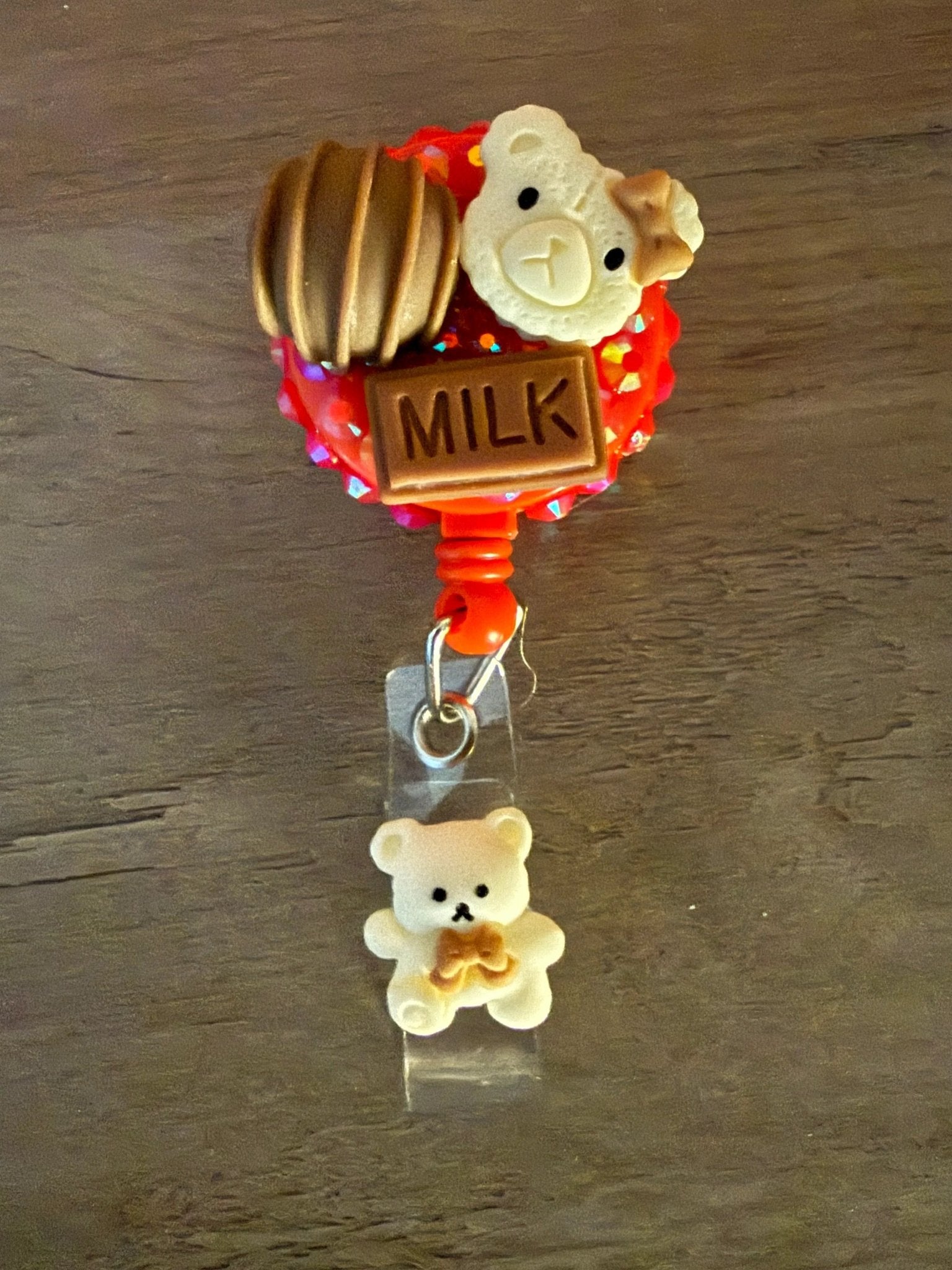 A red badge reel featuring a cute bear charm, a chocolate treat, and a "MILK" sign, with a small cream bear charm below.