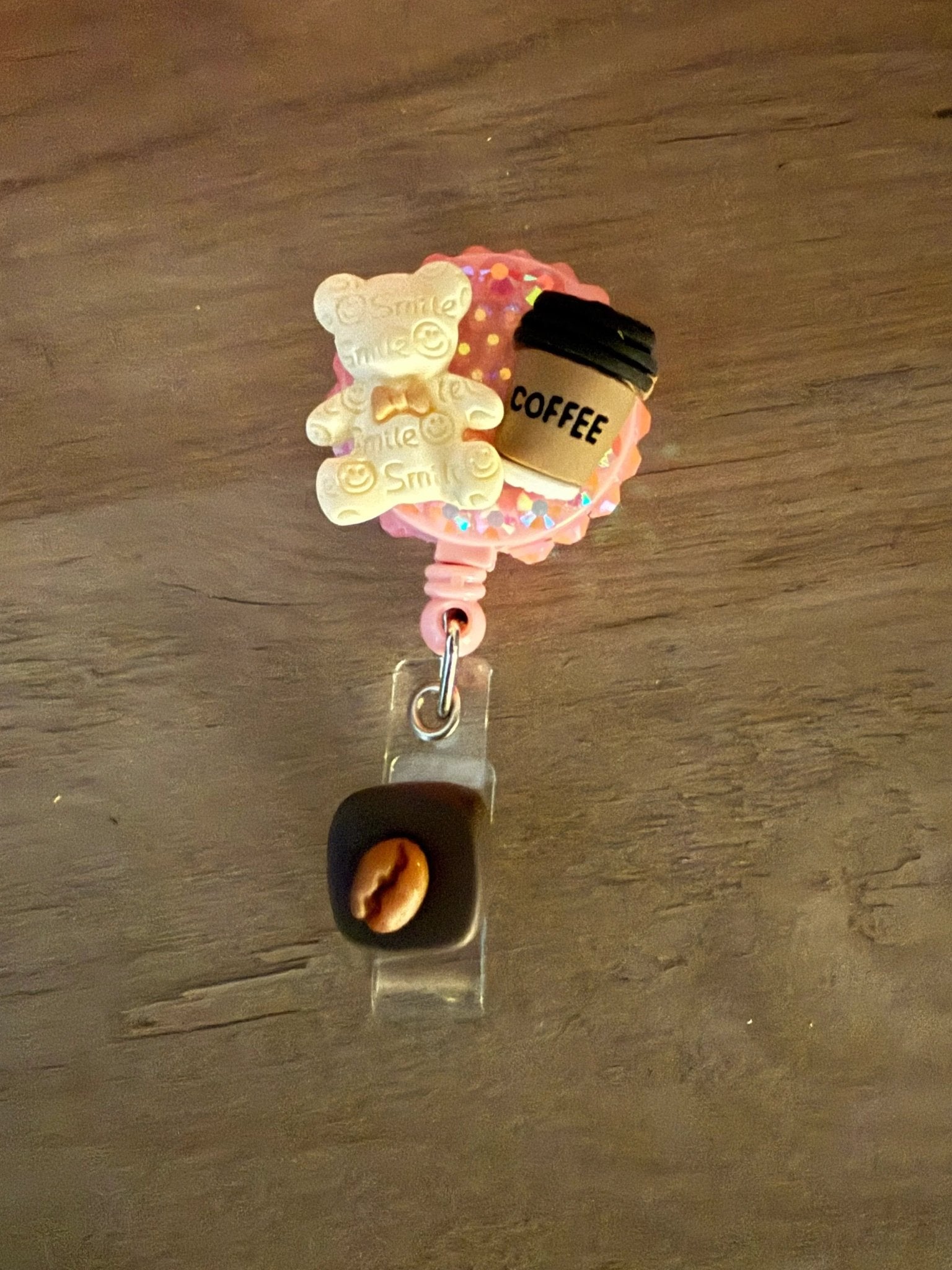 A pink badge reel featuring a smiling teddy bear and a coffee cup charm, with a small coffee bean attached below.