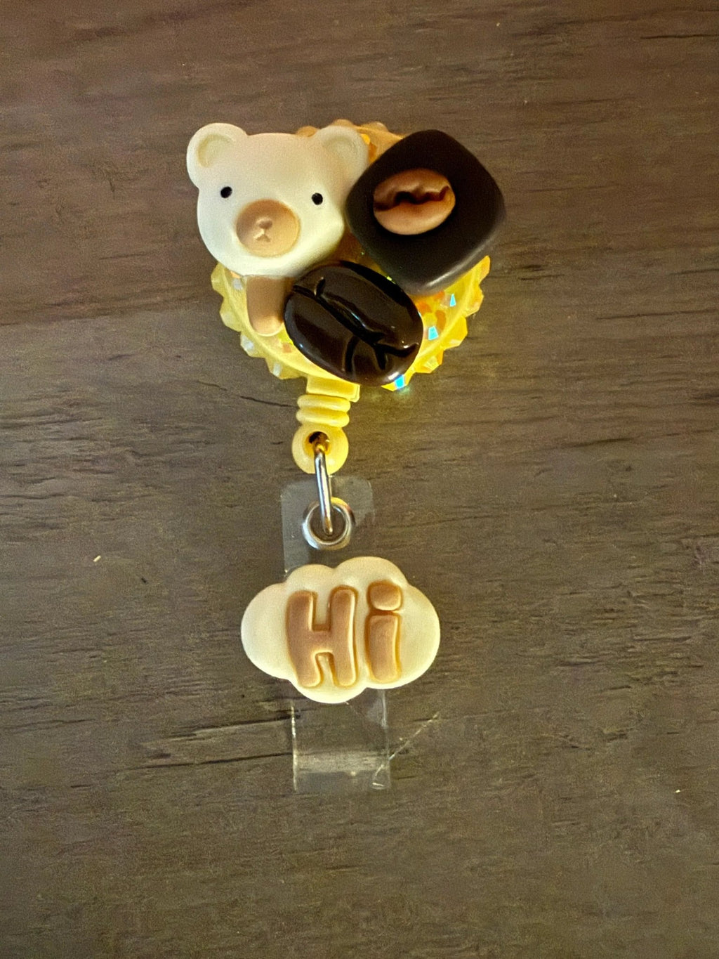 A yellow badge reel featuring a cute bear charm, two coffee bean charms, and a cloud-shaped charm with "Hi" in brown letters.