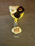 A yellow badge reel featuring a cute bear charm, two coffee bean charms, and a cloud-shaped charm with "Hi" in brown letters.