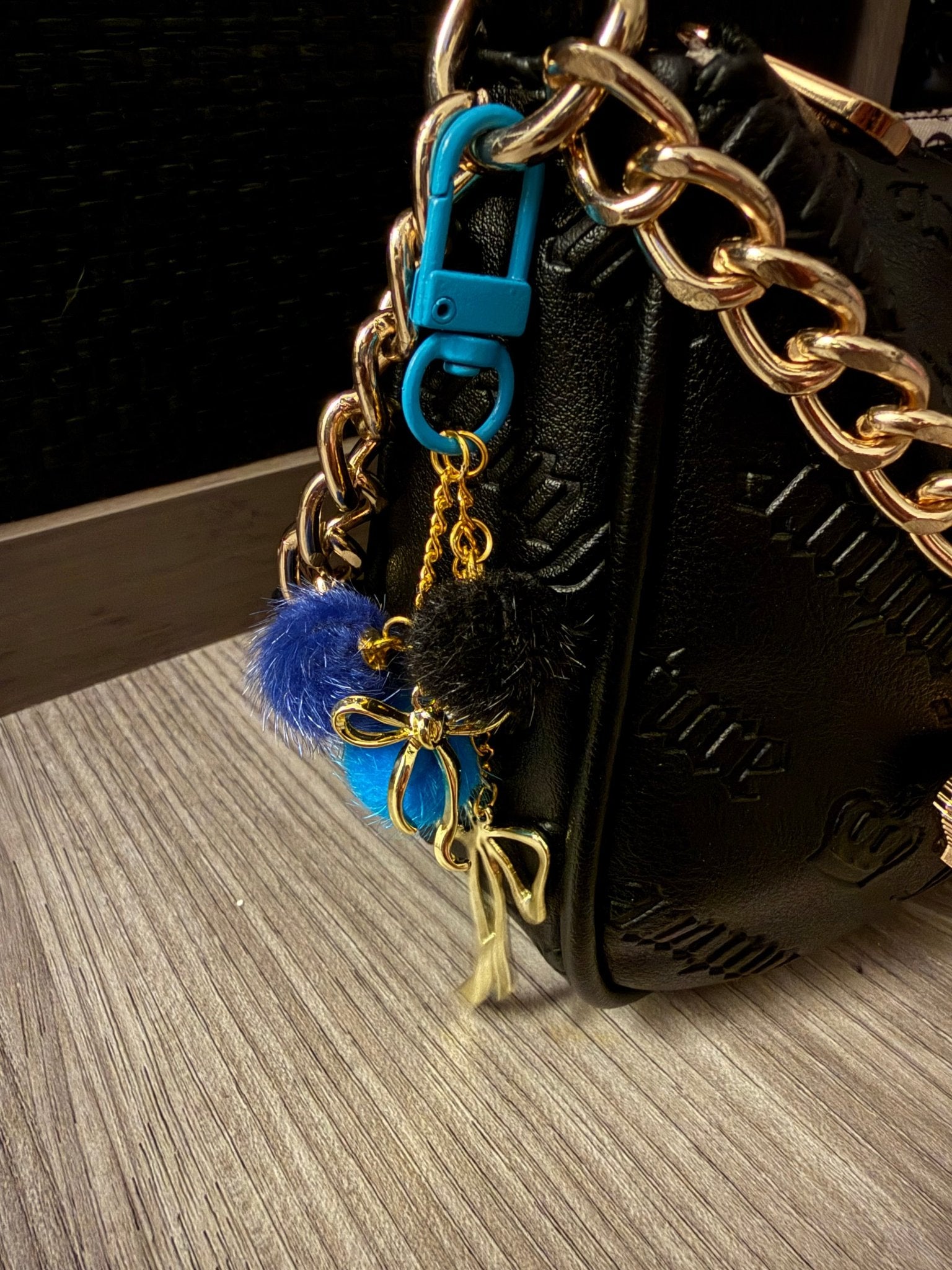 Handmade blue and black pom-pom bag charm with gold bows, shown clipped to a black purse with a gold chain.